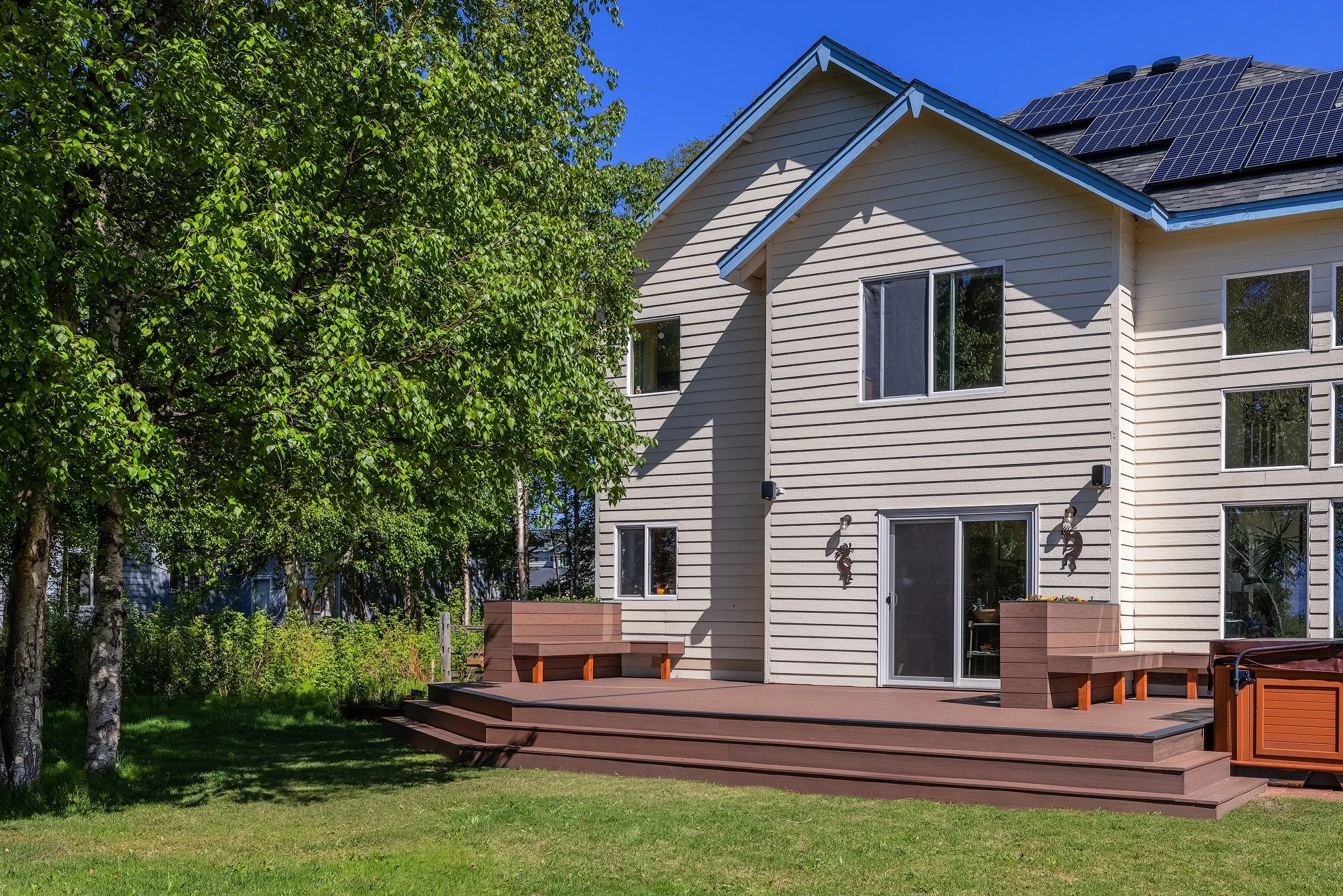 View of the backyard of a house with a spacious composite deck, sliding glass door, multiple windows, and solar panels on the roof. There's a large green tree on the left and a grassy lawn in the foreground.