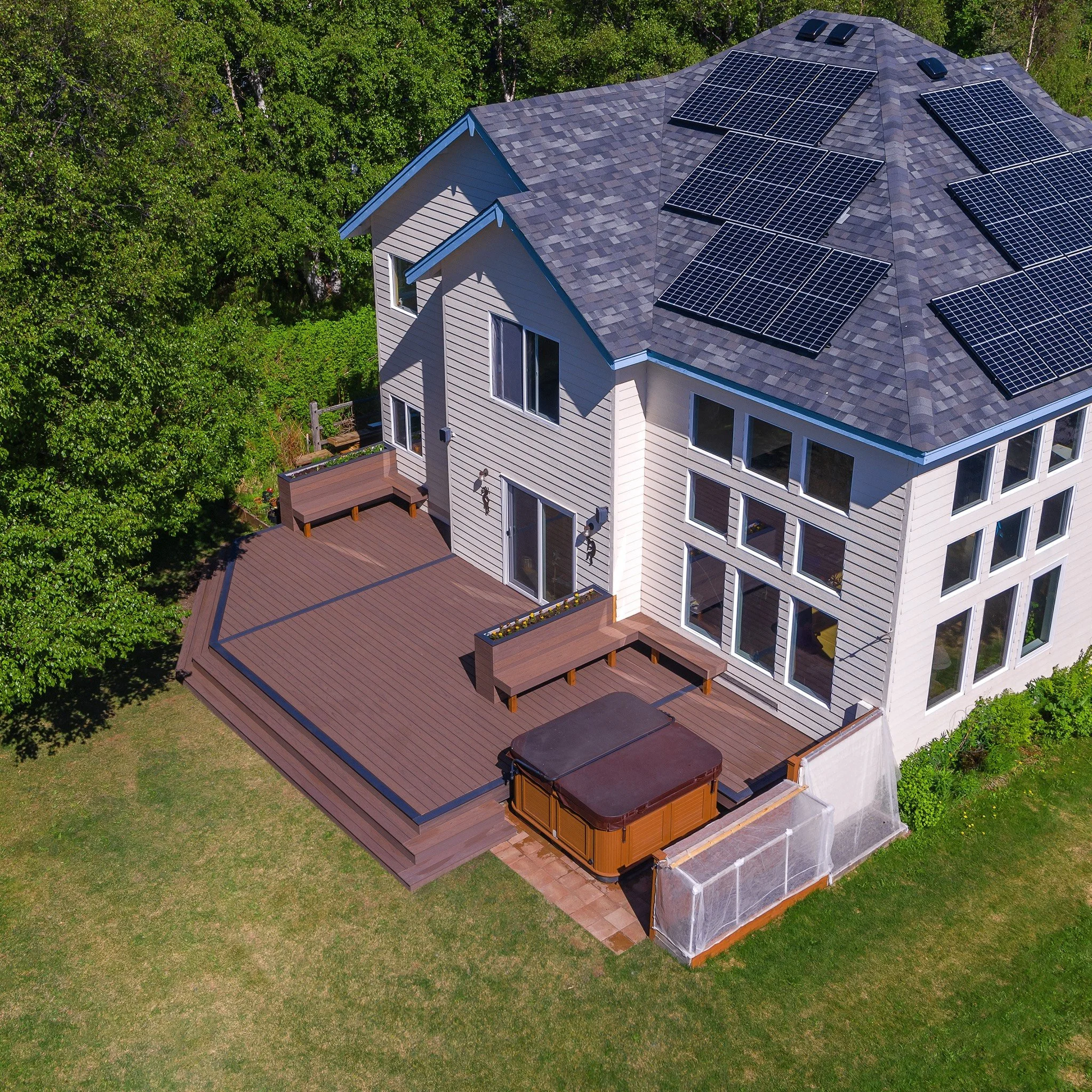 Aerial view of a two-story house with a large rooftop covered in solar panels, a spacious wooden deck, a hot tub, and surrounded by lush green trees.
