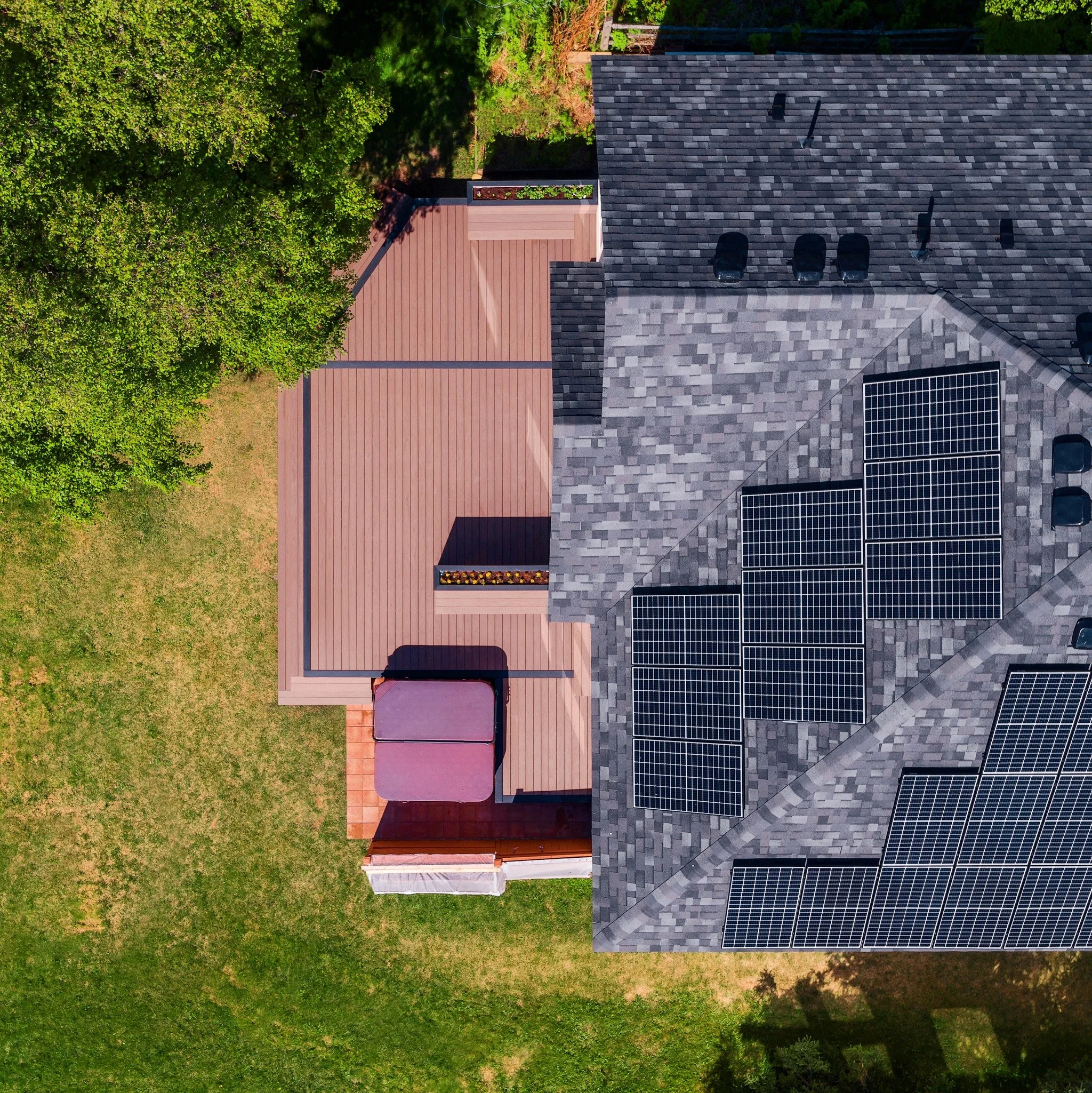 Aerial view of a house with solar panels on the roof, a wooden deck area, green lawn, and surrounding trees.