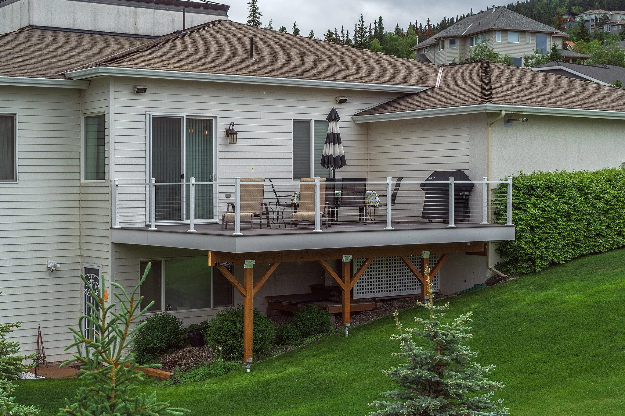 A raised composite deck attached to the backyard of a house, with outdoor furniture, an umbrella, and a barbecue grill, overlooking a green lawn and surrounded by shrubs and trees.