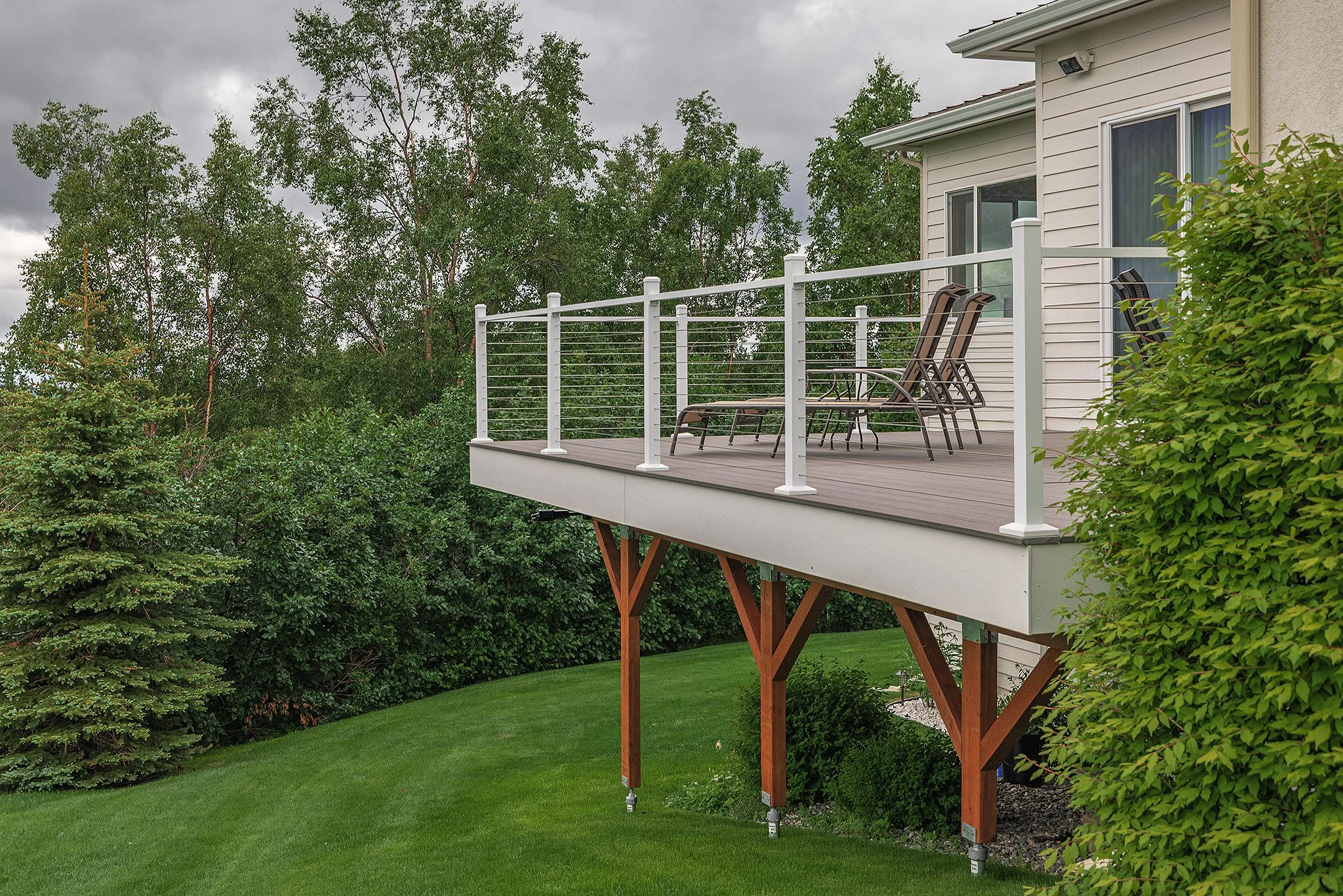 A composite deck with white railings extends from a house, with a grassy yard and trees in the background, and three lounge chairs on the deck