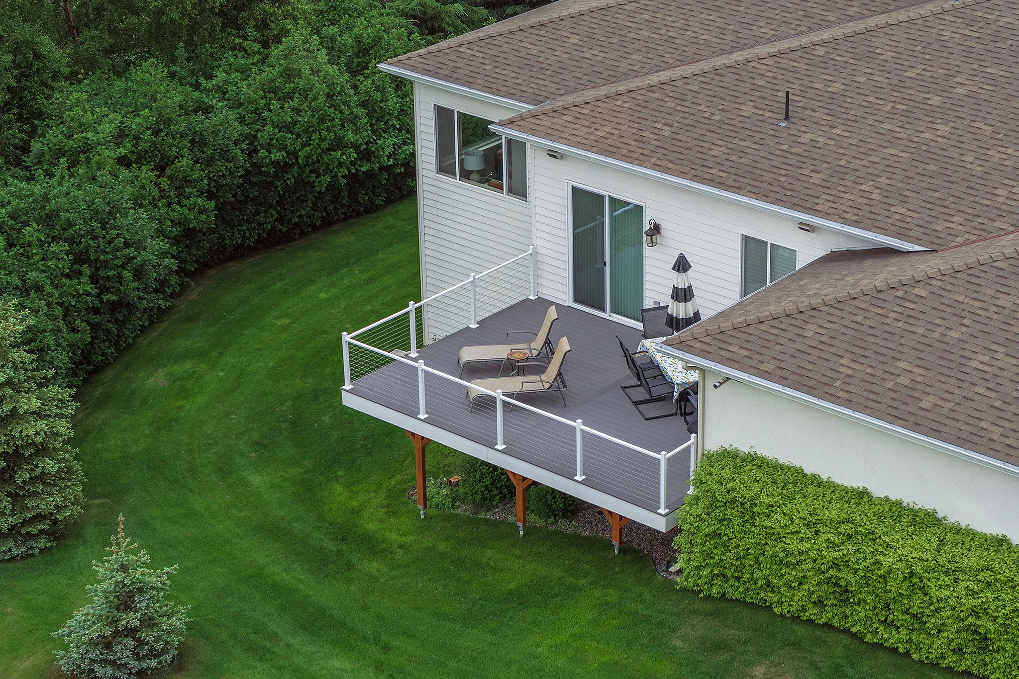 Aerial view of a backyard patio with outdoor furniture, including lounge chairs, a table with a tablecloth, and an umbrella, attached to a white house with sliding glass doors, surrounded by green grass and trees.