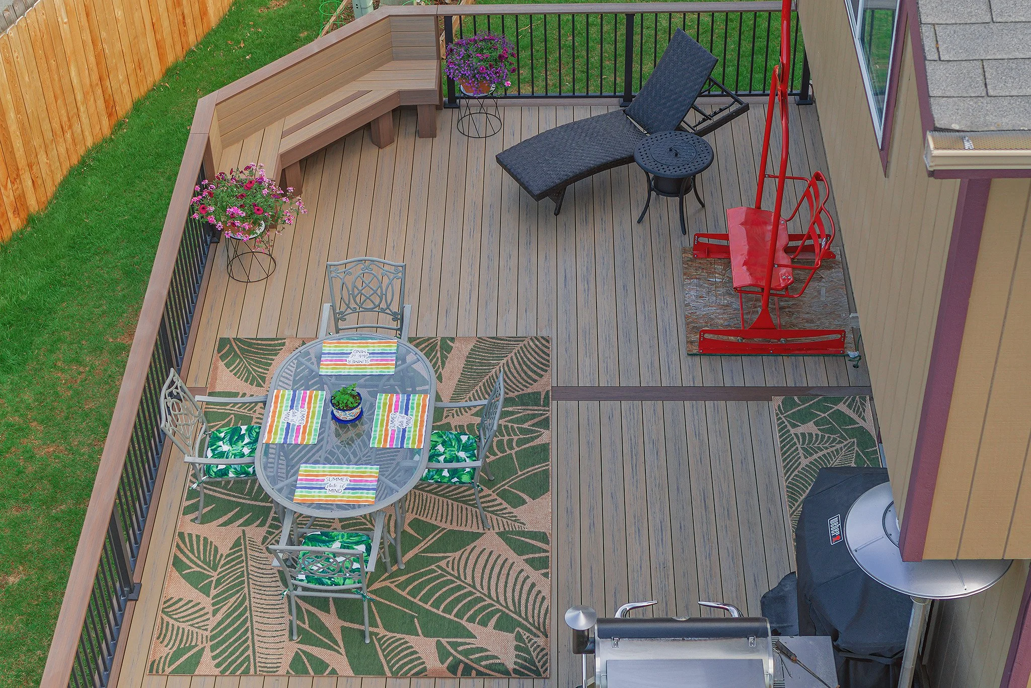 An outdoor composite deck with furniture, including a dining table with four chairs, knives, and a potted plant, a chaise lounge, a small round side table, and a red sled and a black grill cover in the corner.