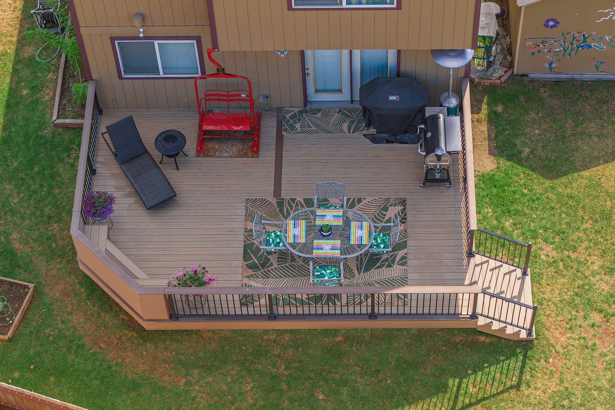 A backyard deck with outdoor furniture, a grill, a lounge chair, and decorative plants surrounded by a grassy yard.