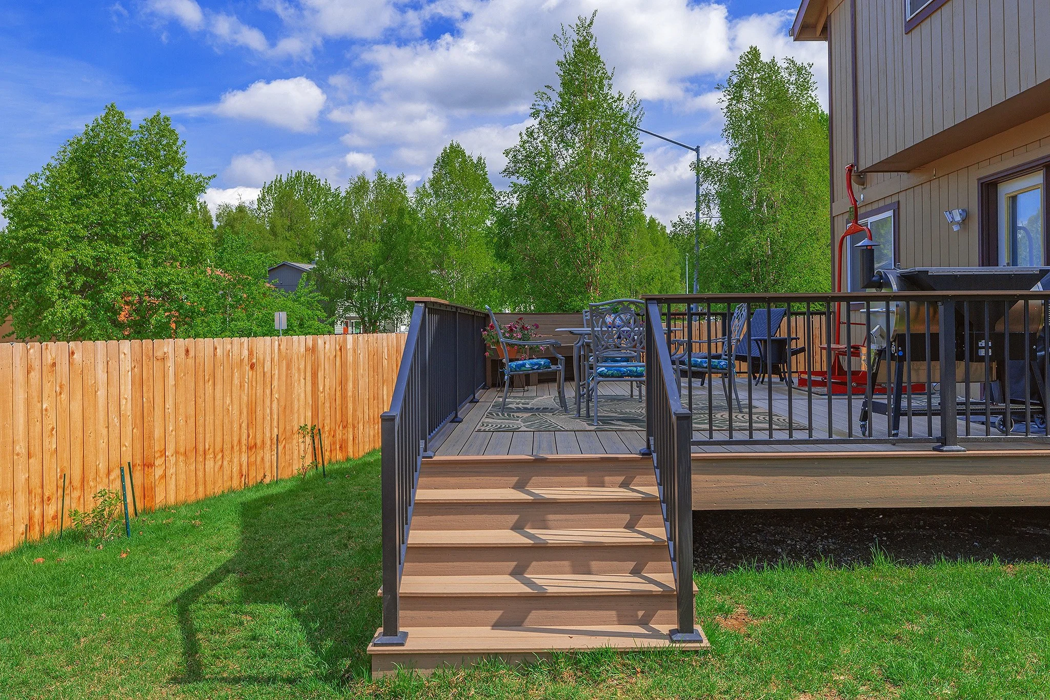 Composite deck with outdoor furniture and garden equipment, overlooking a fenced yard with green trees under a partly cloudy sky.