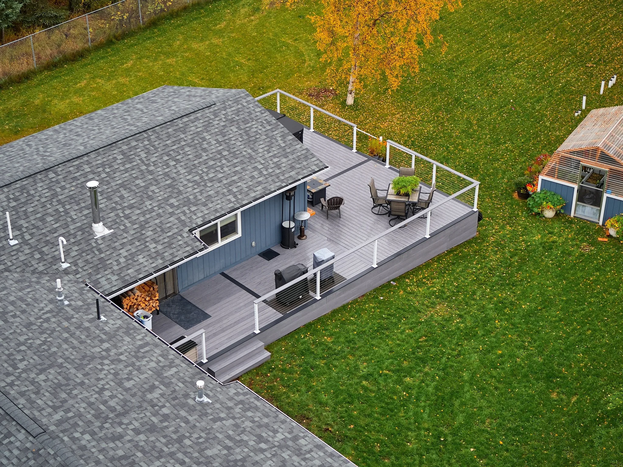 A house with an attached outdoor deck, outdoor furniture, and a backyard with grass and trees, shown from an aerial view.