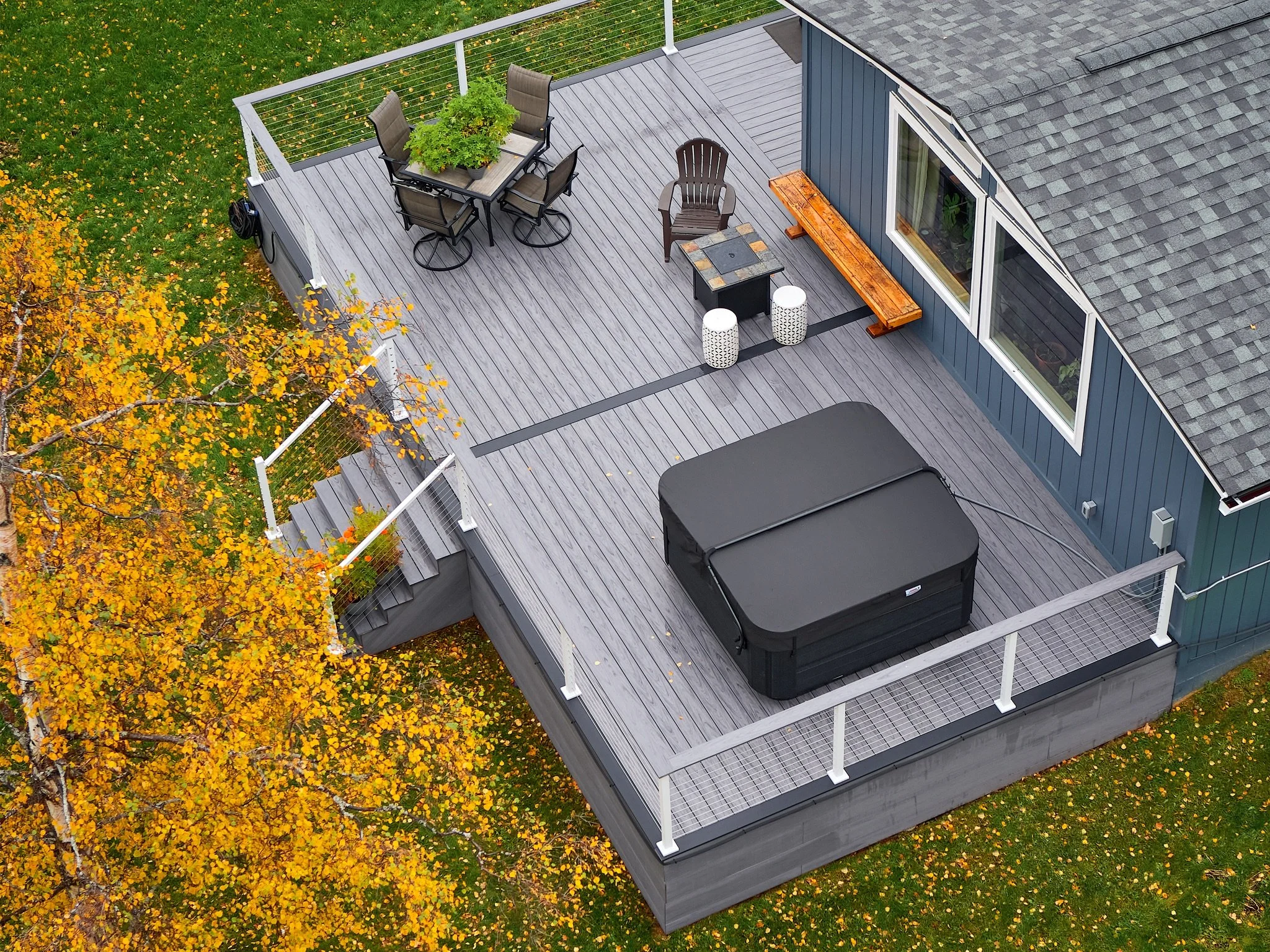 Aerial view of a large outdoor deck attached to a house, with a hot tub, outdoor dining set, and various seating options, surrounded by green grass and trees with yellow and orange autumn leaves.