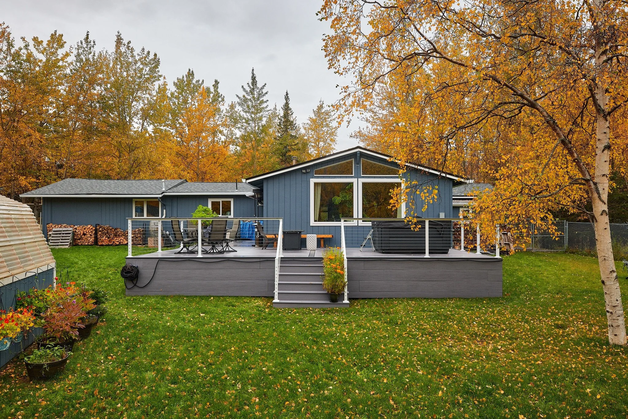 A house with a large deck overlooking a yard with autumn trees, lawn, potted plants, and outdoor furniture.