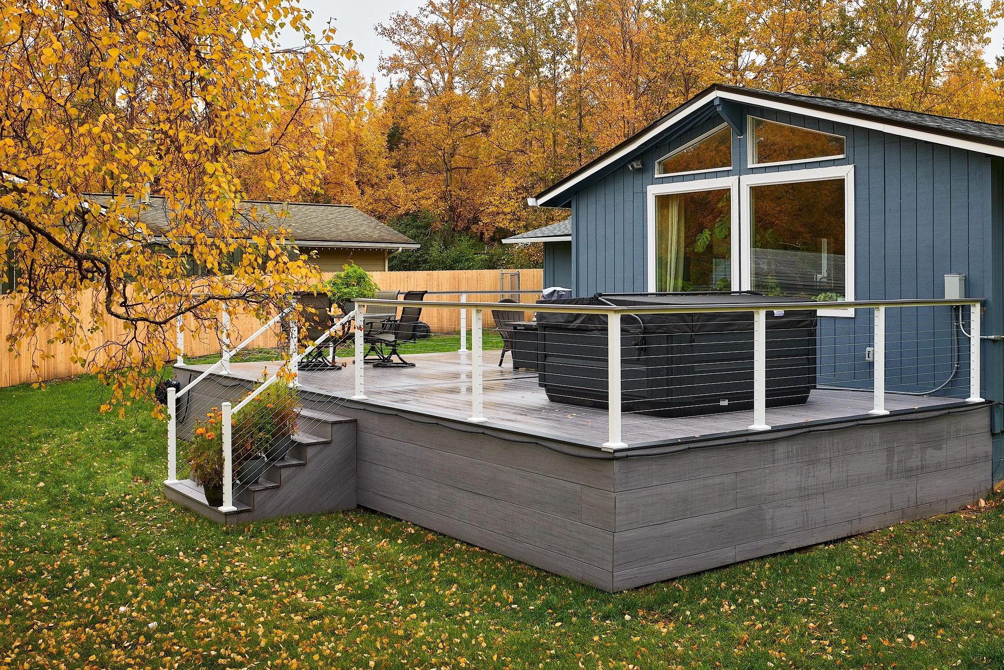 A backyard with a composite deck featuring outdoor furniture, a hot tub, and a black grill, surrounded by green grass and autumn trees.