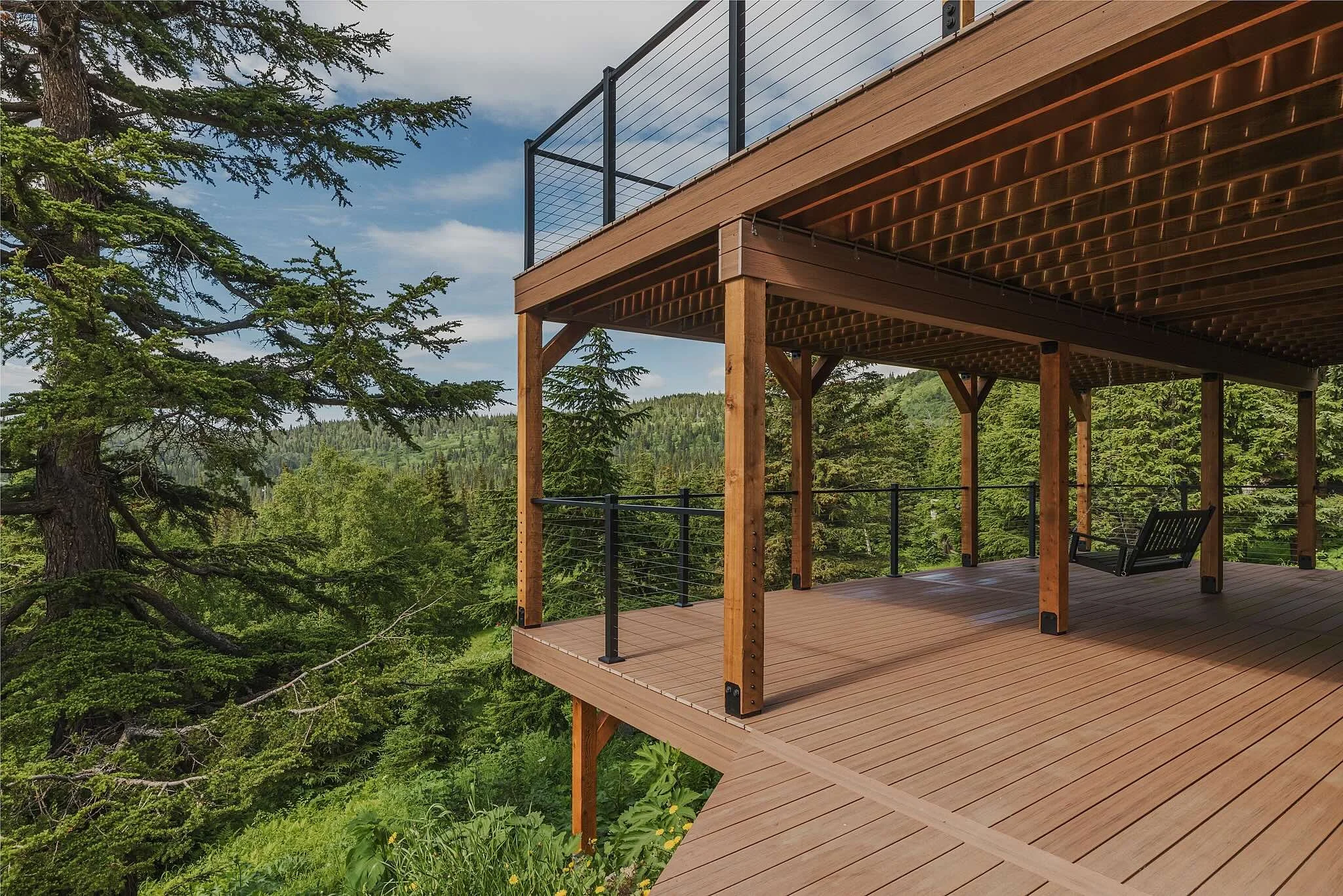 Composite deck with a black metal railing, overlooking a forested landscape with tall trees and hills in the background, under a partly cloudy sky.