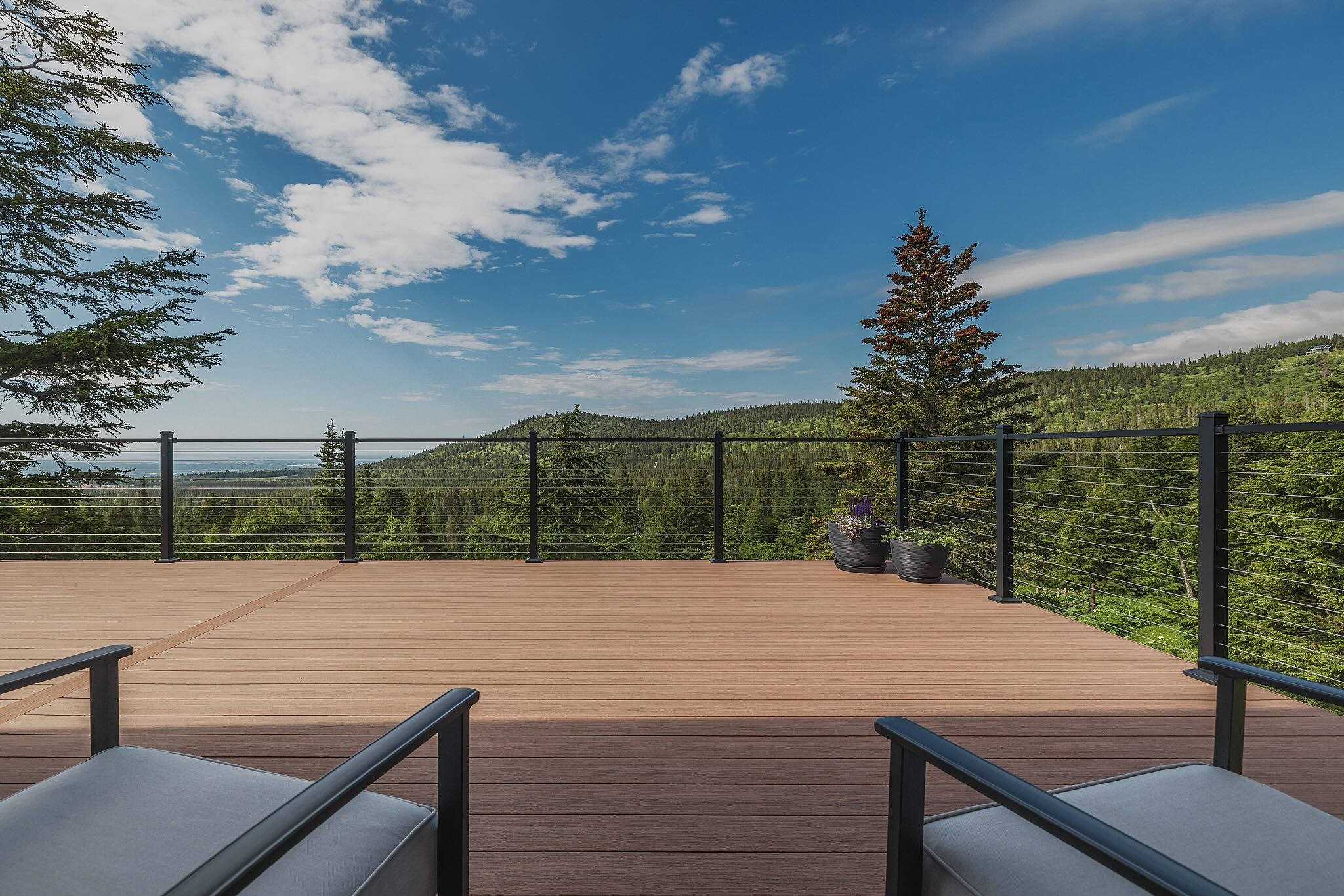 View from a composite deck with black railing overlooking a forested mountain landscape under a blue sky with white clouds. Two gray chairs with black armrests are placed on the deck, and there are two potted plants on the right side.