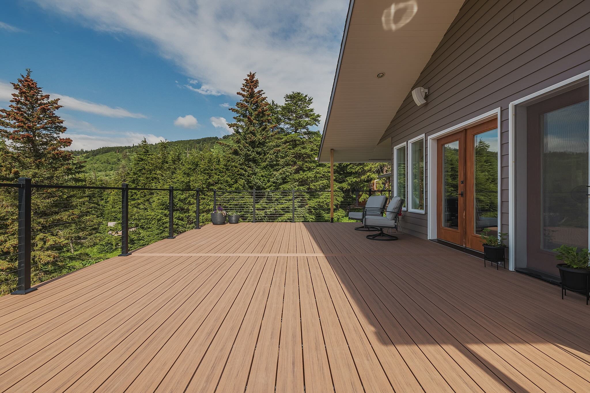 Large comoposite deck with patio chairs, potted plants, and house exterior with windows and a glass door, surrounded by green trees and hills under a partly cloudy sky.