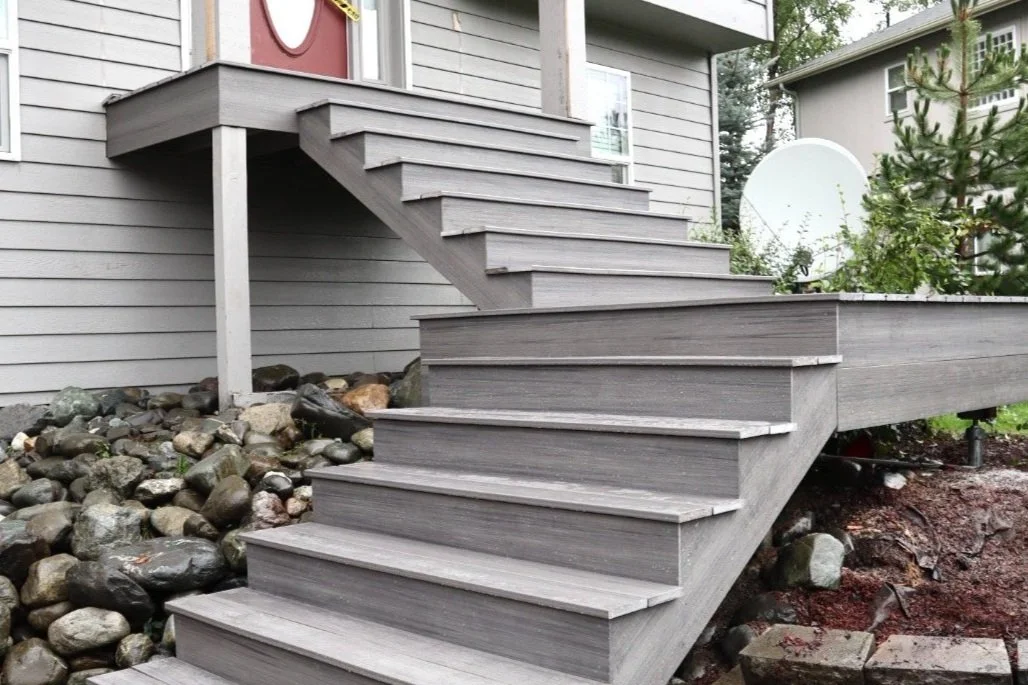 Gray composite outdoor staircase leading to a house deck, with rocks and soil underneath.