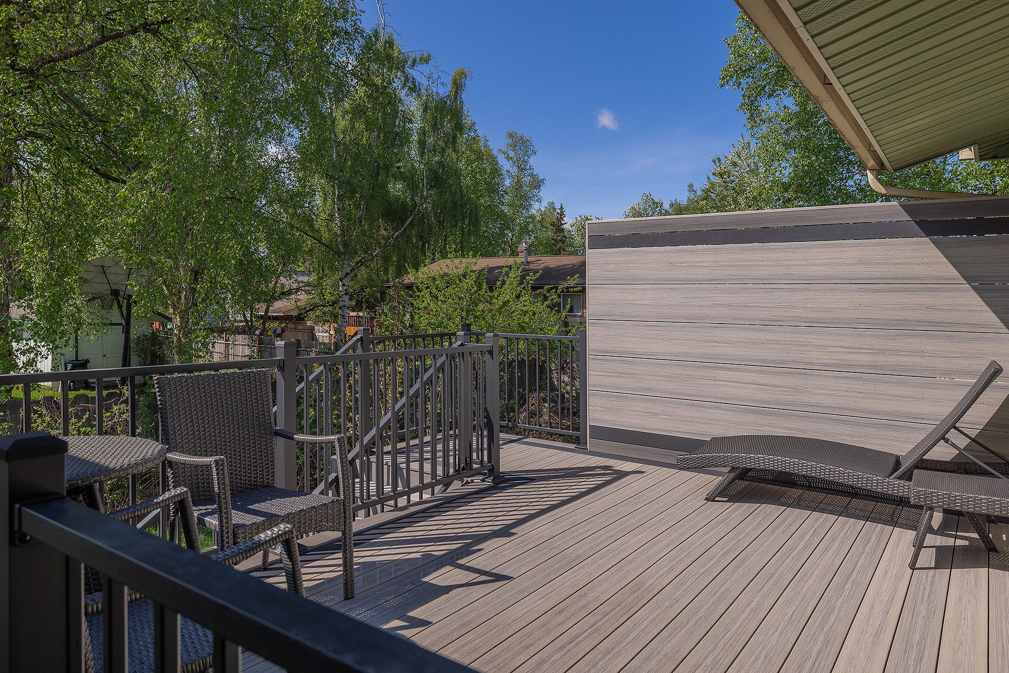 A composite outdoor deck with patio furniture and lounge chairs, surrounded by trees and a railing, under a blue sky with some clouds.