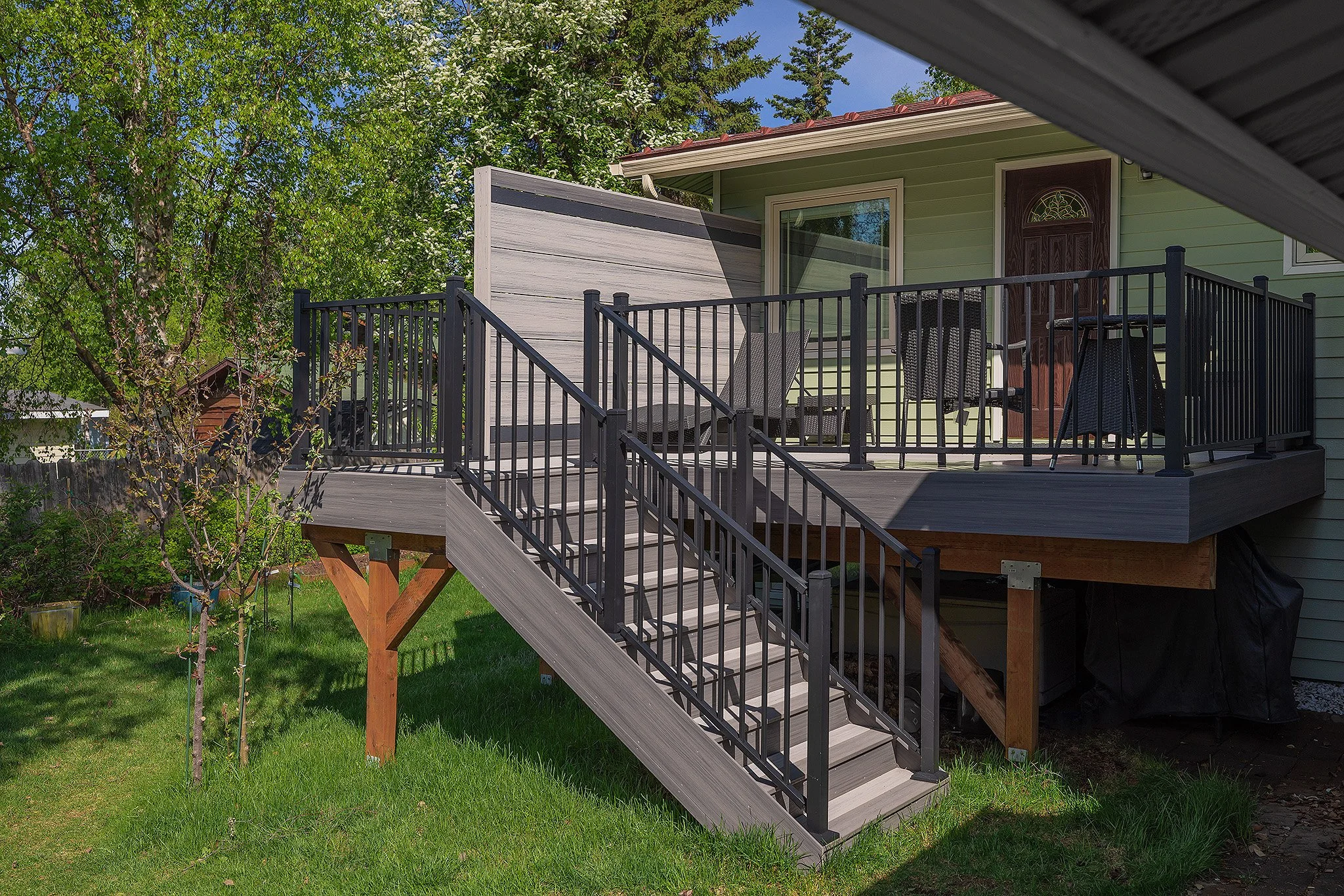 A backyard deck with black aluminum railings and stairs leading down to a green lawn. The deck has outdoor furniture including a table and chairs. Part of the house with a door and window is visible, with trees and greenery in the background.