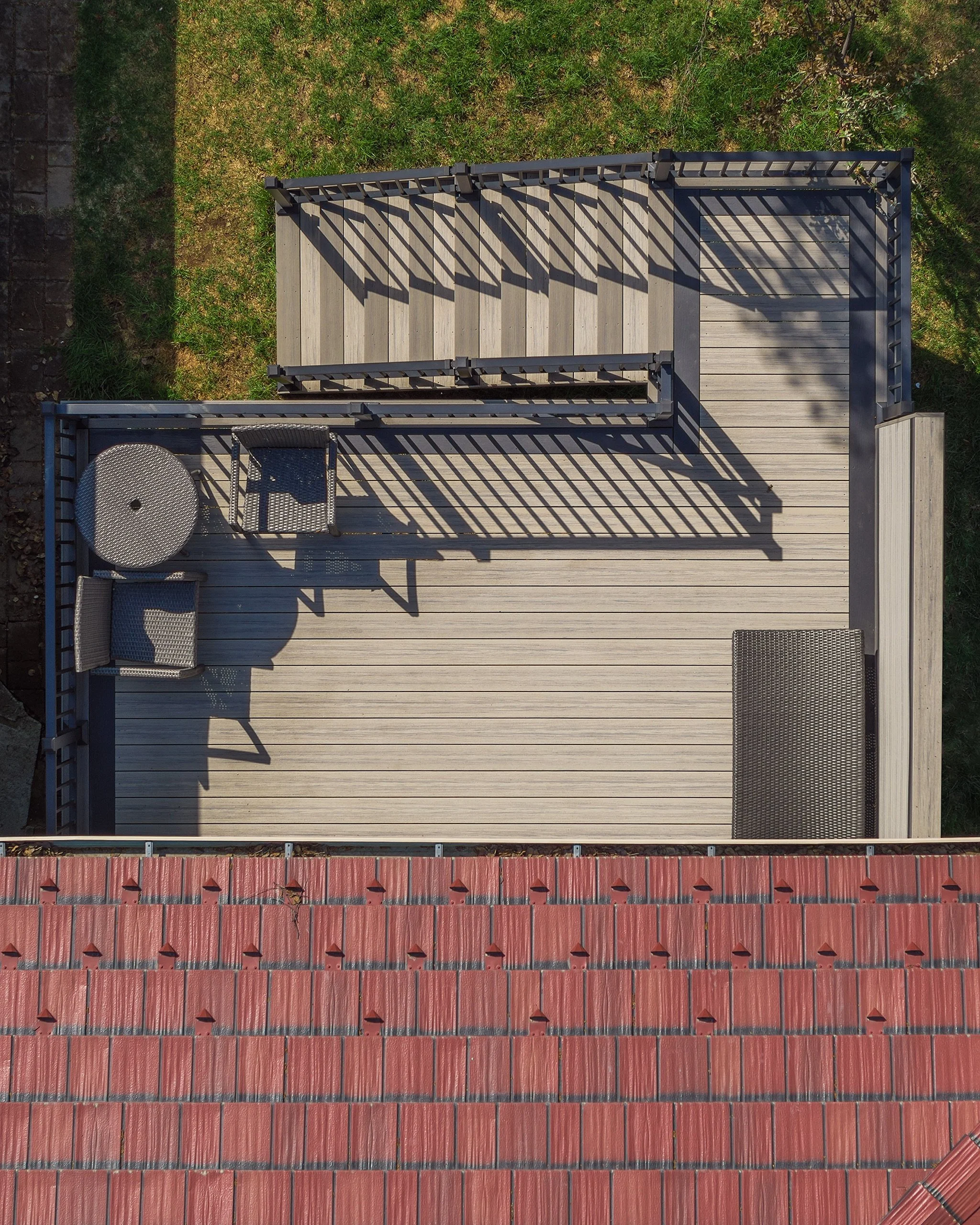An aerial view of a composite outdoor deck with seating and shadows, bordered by a grassy area and a red tiled roof.