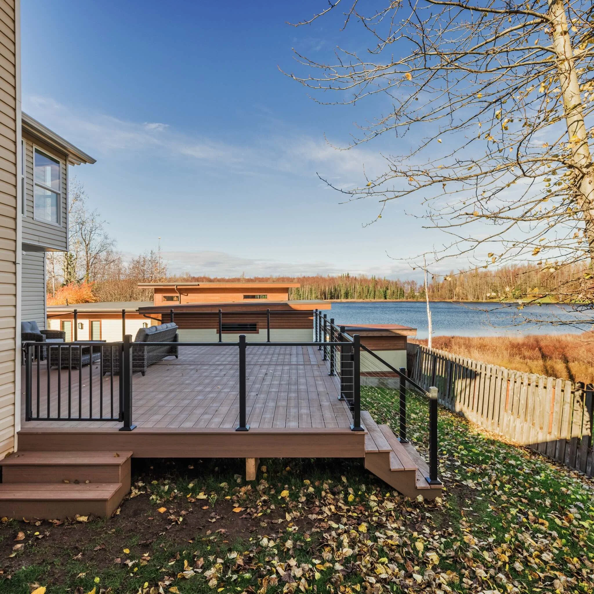 A composite deck with black railings overlooking a lake, with a house partially visible on the left, and trees with fallen autumn leaves in the background.
