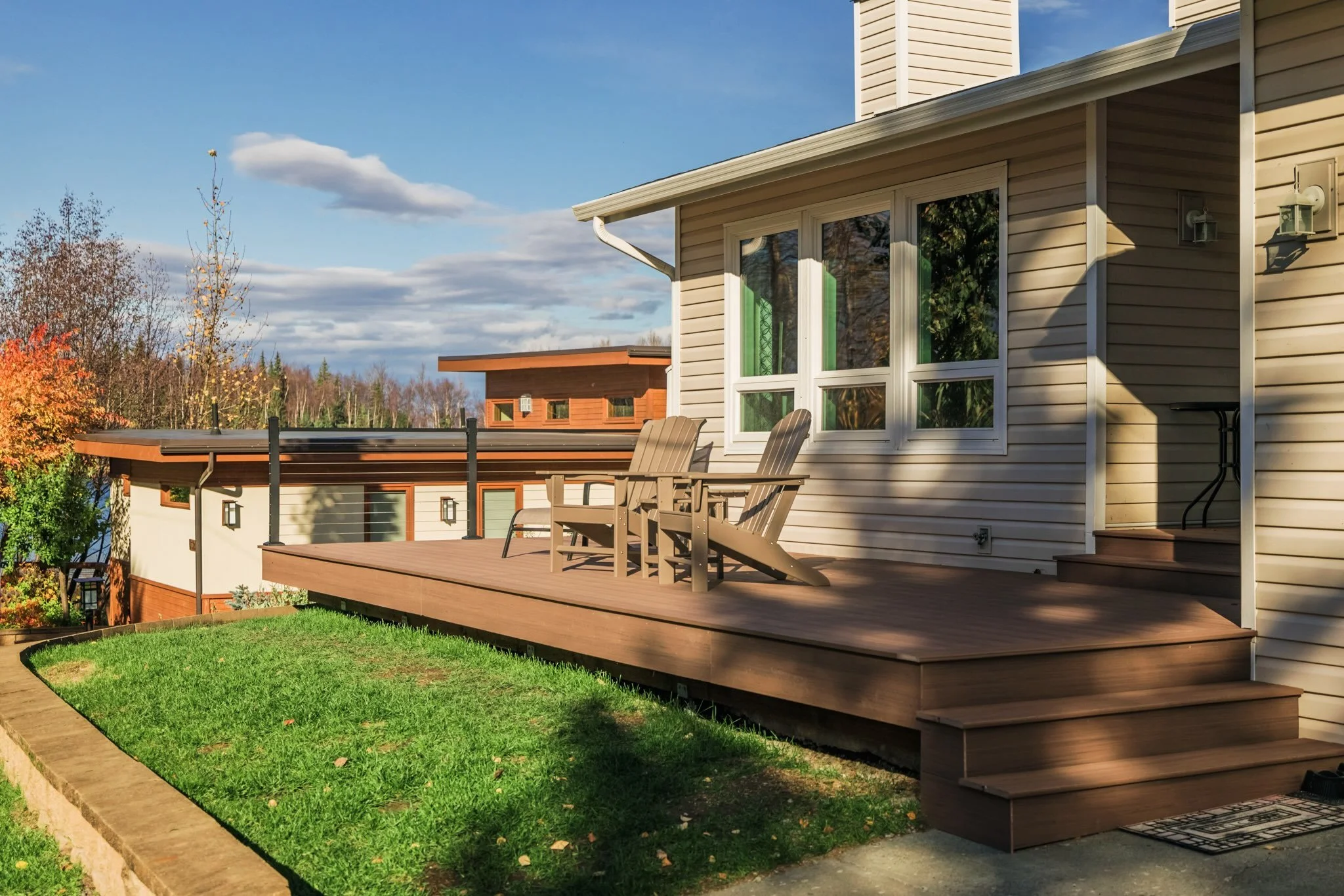 A house with a composite deck and two Adirondack chairs, sliding glass doors, stairs leading up to the deck, and a lawn in the foreground during daytime.