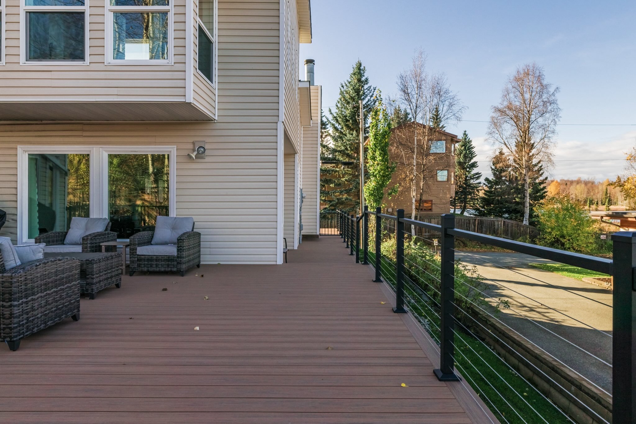 Luxury outdoor balcony with wicker furniture, including chairs and cushions, overlooking a neighborhood with trees and multi-story houses on a sunny day.
