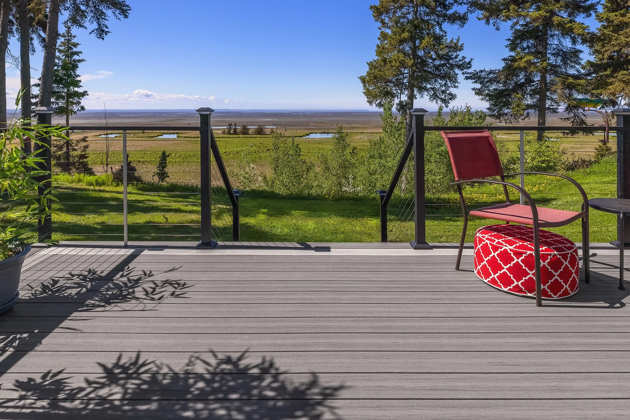 A composite deck with two chairs, one red and one with red and white pattern, overlooking a green landscape with trees, grass, and small bodies of water under a blue sky.