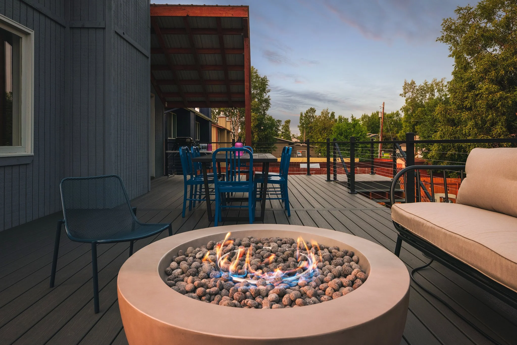 Outdoor second-story deck with seating area, including a fire pit, table with chairs, and a lounge chair, surrounded by trees and a railing, during sunset or late afternoon.
