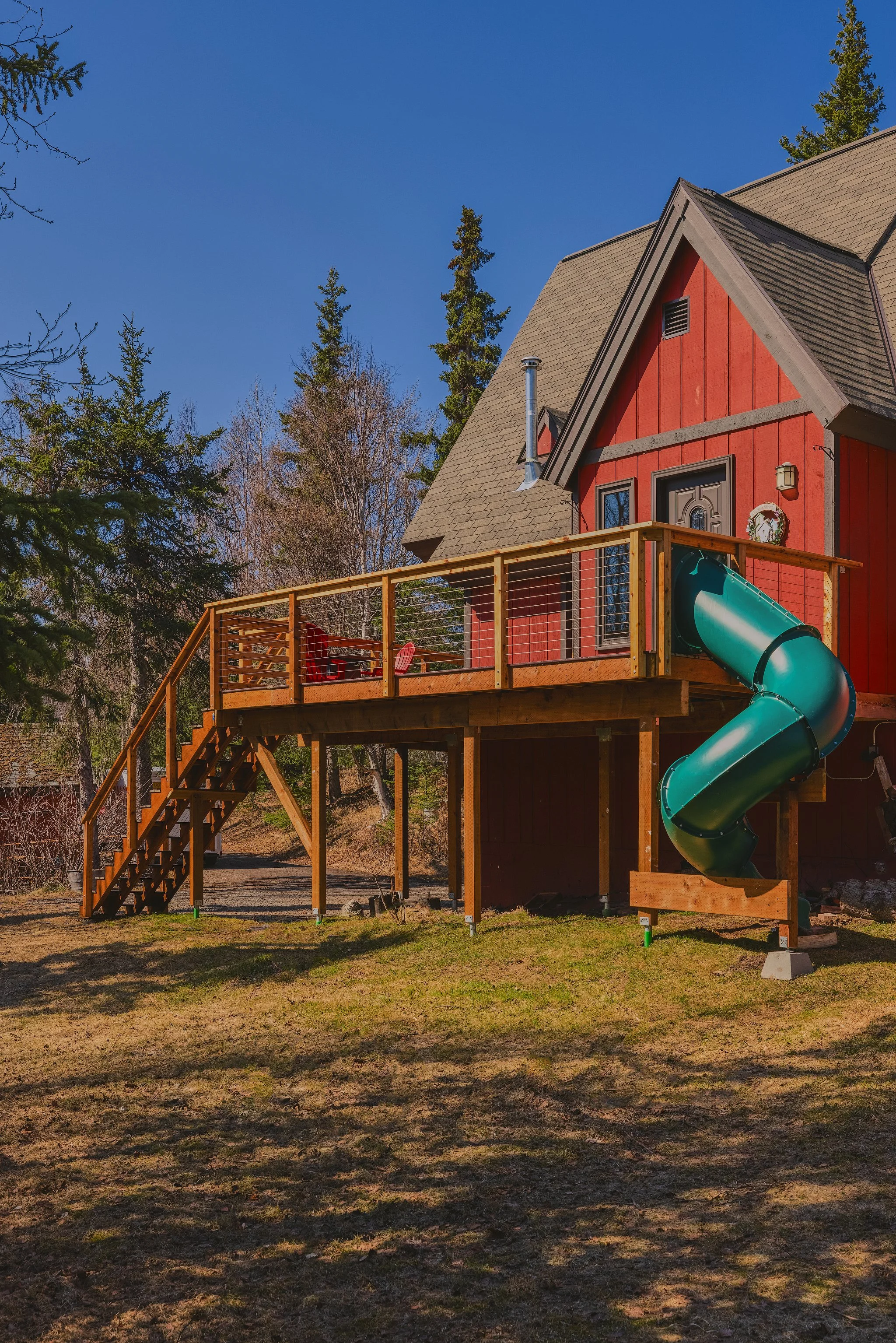 Red house with a green slide attached to a wooden deck in a backyard, with trees and a blue sky in the background.