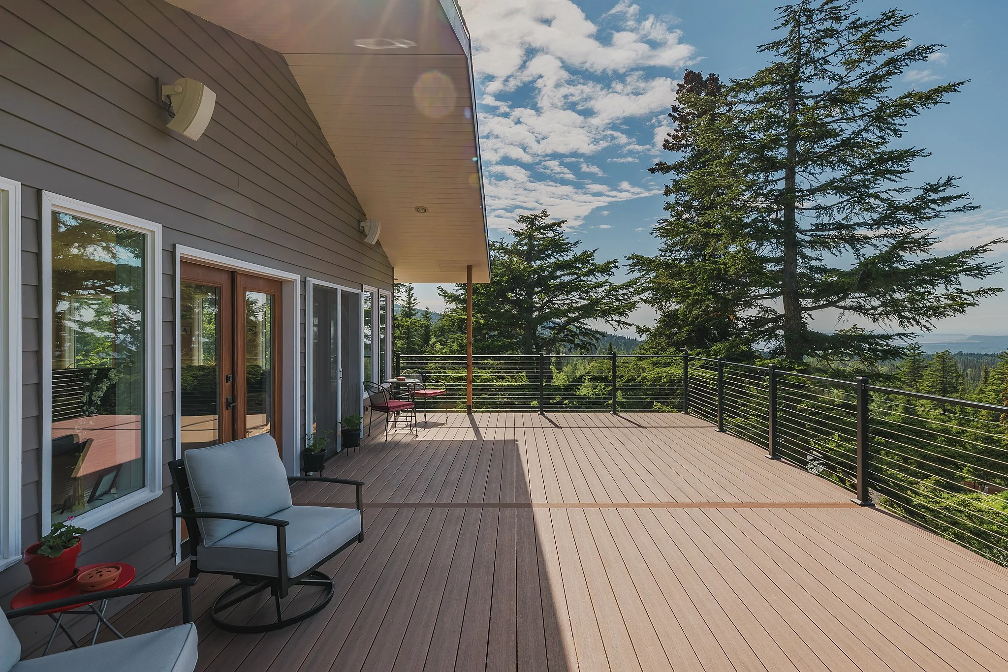 Large outdoor balcony with composite decking, black metal railing, and patio furniture, overlooking a green forest with tall trees under a partly cloudy sky.