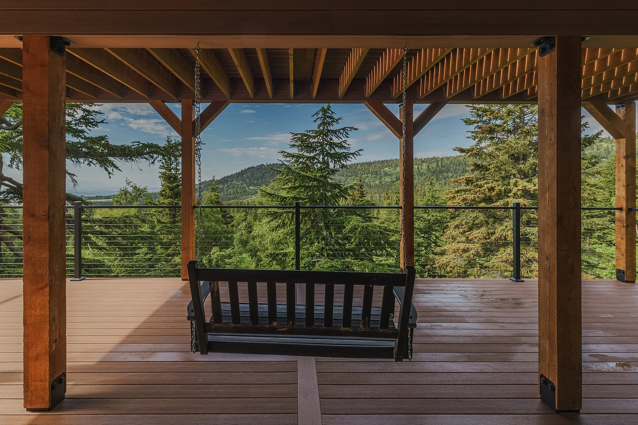 A composite decking porch with a black swing, overlooking a lush green forest and distant mountain under a partly cloudy sky.