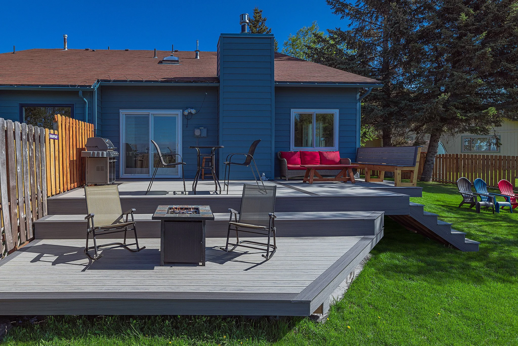 Backyard patio with outdoor furniture, including chairs, a fire pit, a barbecue grill, and a red outdoor couch, surrounded by a wooden fence and green grass, with a blue house in the background under a clear sky.