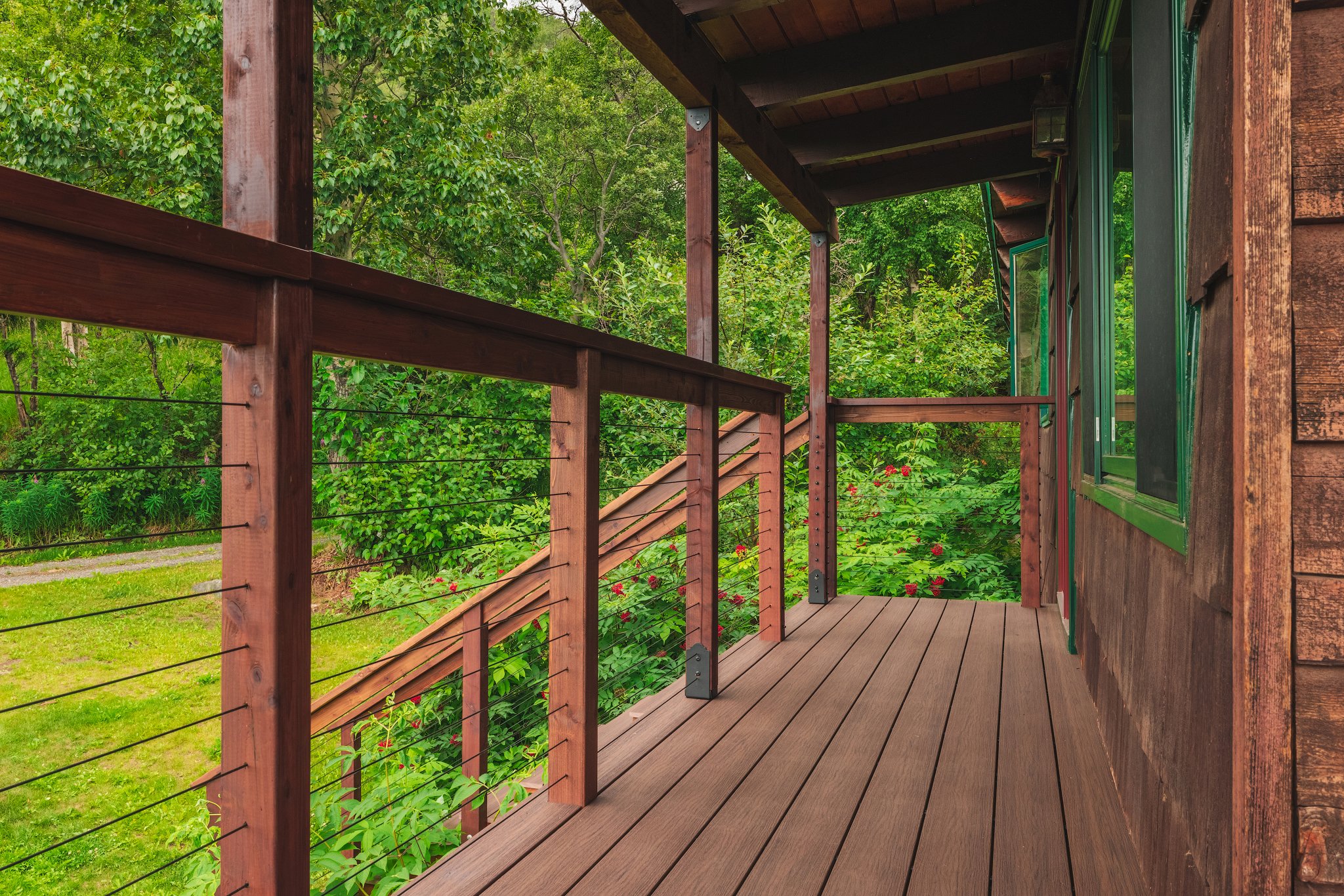 A composite decking balcony with a railing overlooking a lush green backyard with trees and flowering bushes.