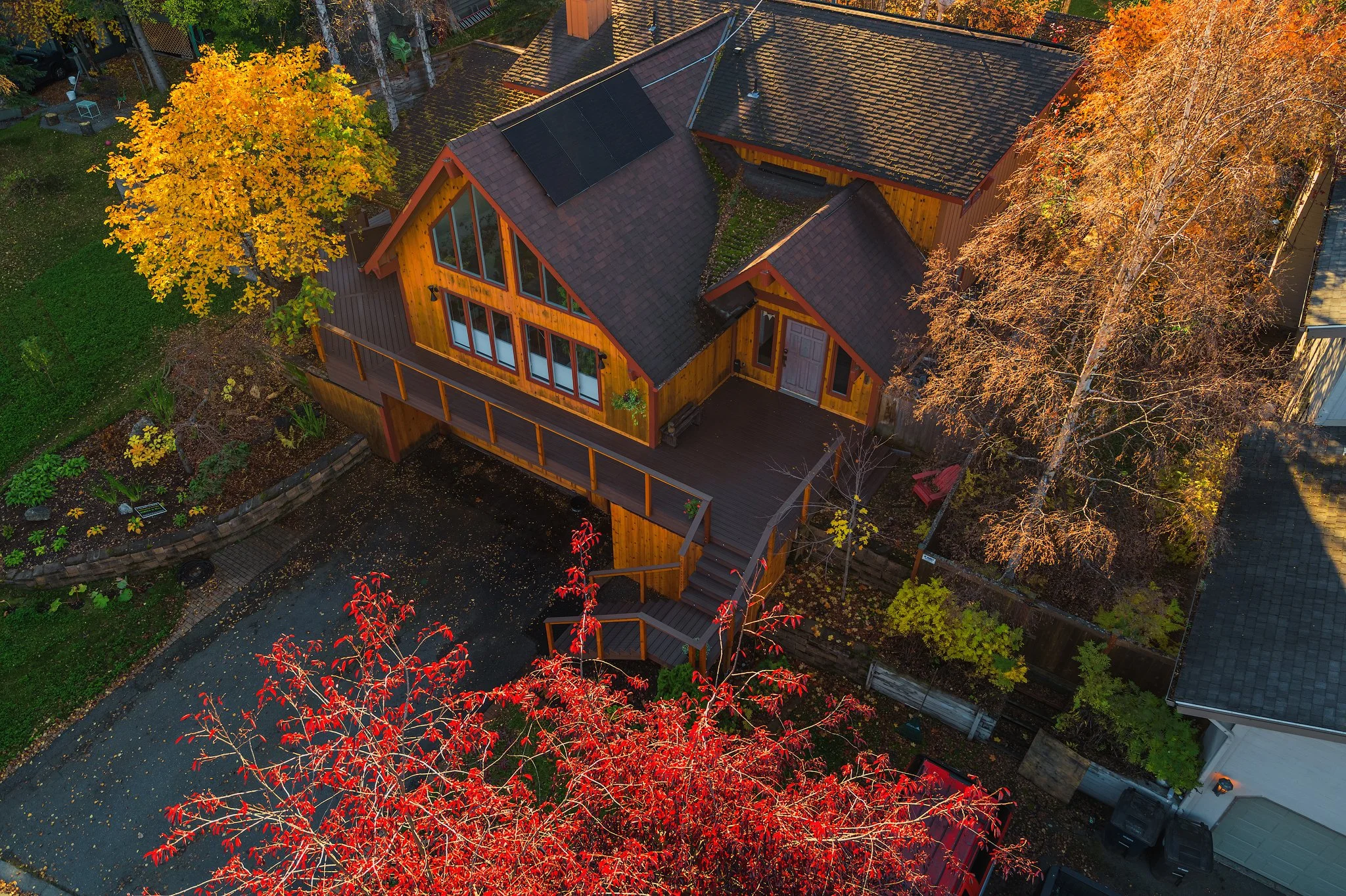 Aerial view of a wooden house with solar panels on the roof, surrounded by trees with autumn foliage, a deck with stairs, and a driveway.