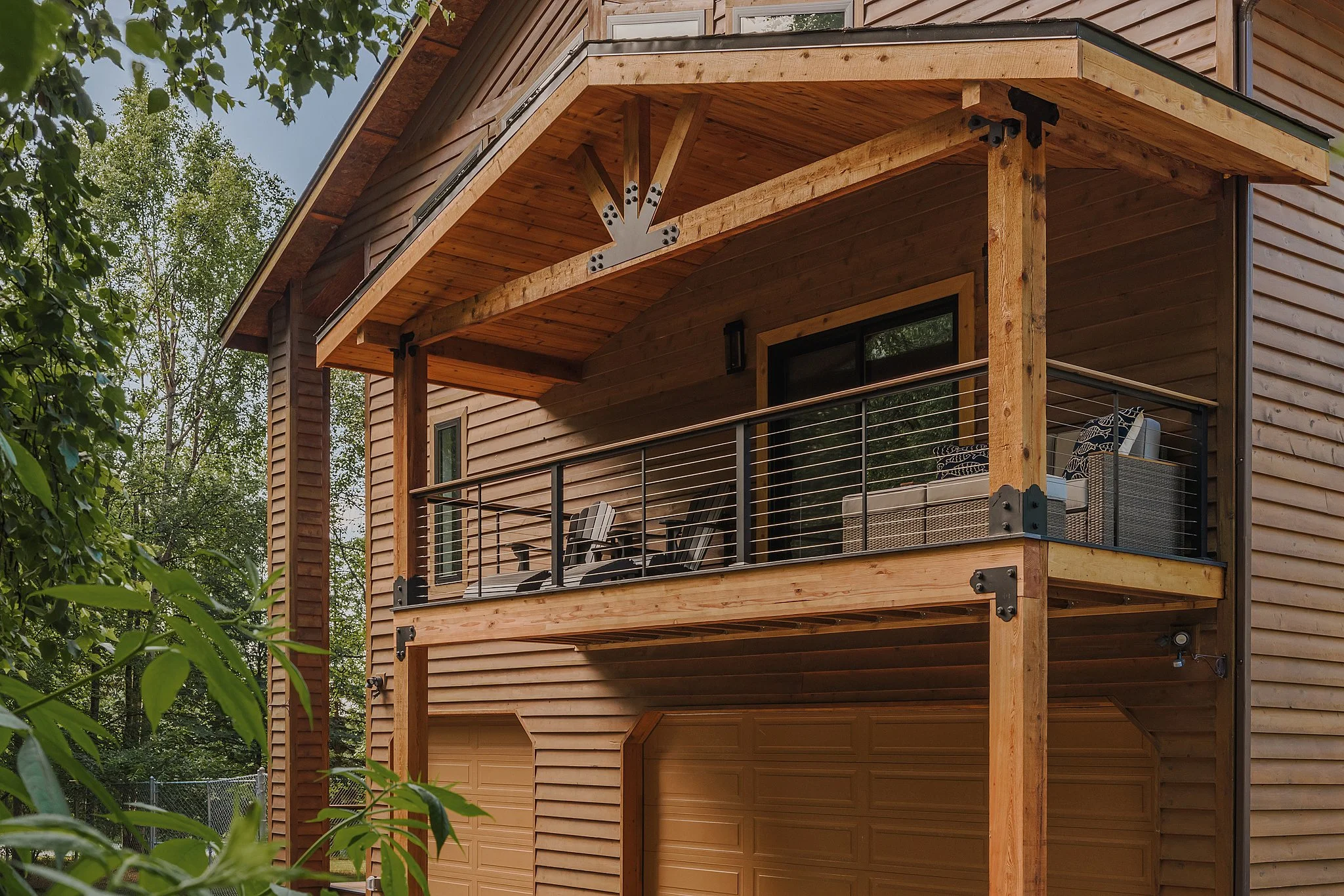 A two-story wooden house with a balcony on the second floor with black metal railings, outdoor furniture, and a neighboring house in a wooded area.