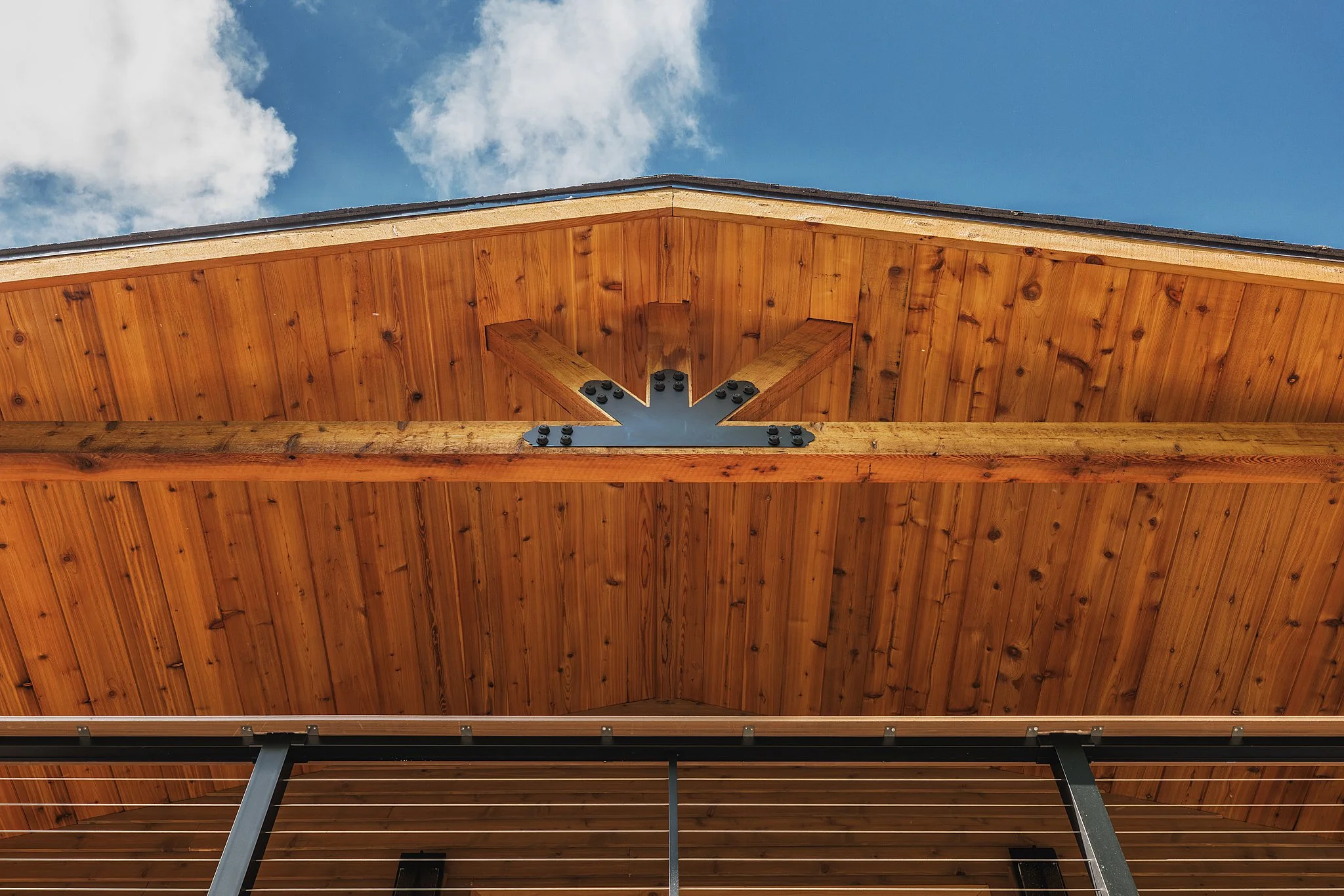 Looking up at the underside of a wooden roof structure with metal braces, against a background of blue sky and clouds.