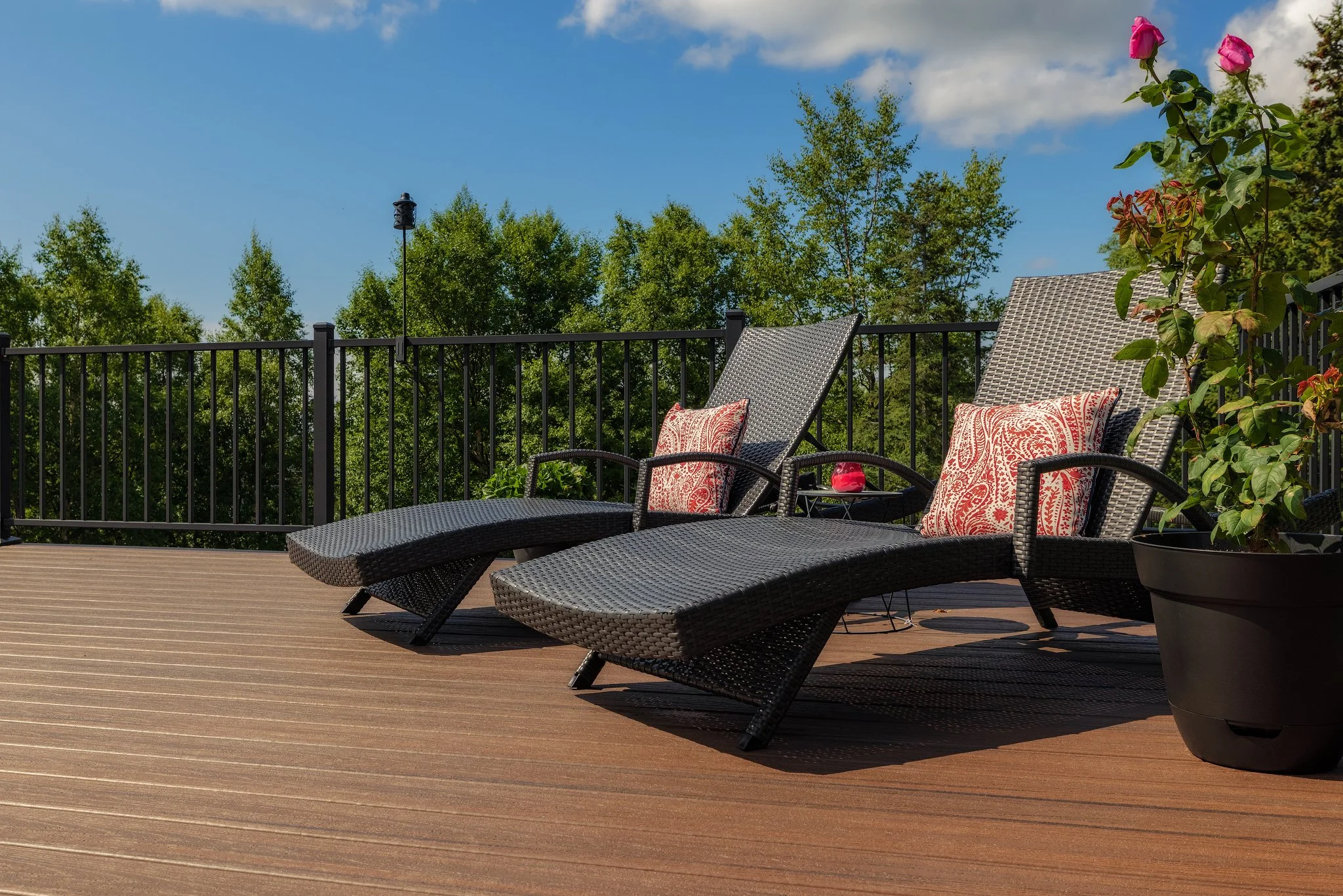 Two black wicker lounge chairs with decorative pillows on a trex deck, next to a potted plant, with trees and a black aluminum metal railing in the background under a blue sky.