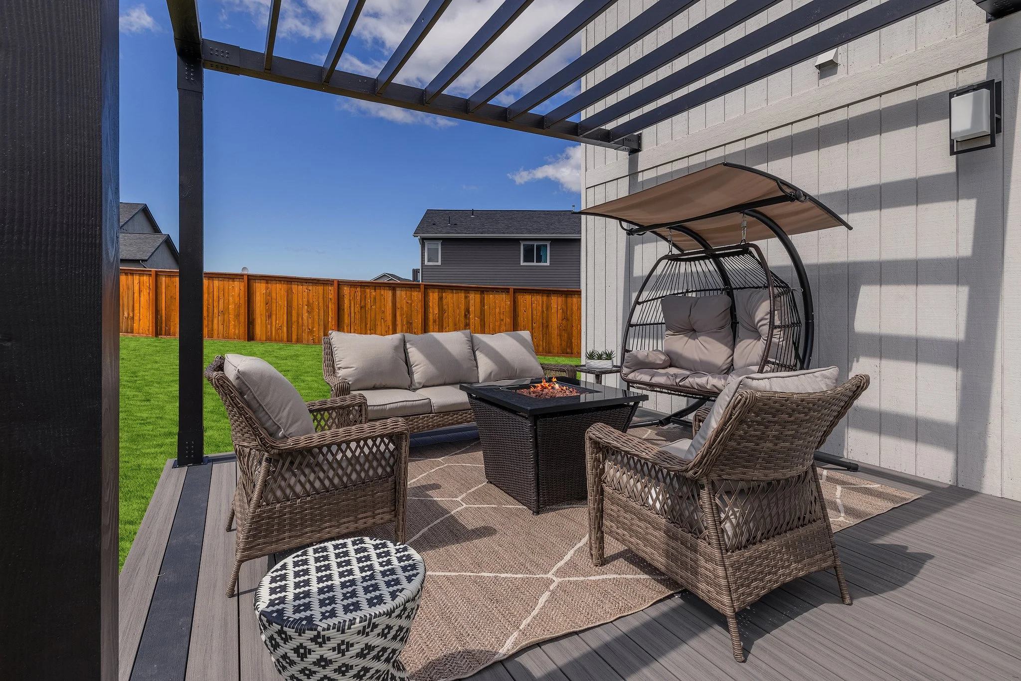 Outdoor patio with seating area, including wicker chairs, a cushioned loveseat, a hanging egg chair, a firepit table, a patterned pouf, and a rug, under a pergola with a wooden fence in the background and a clear blue sky.