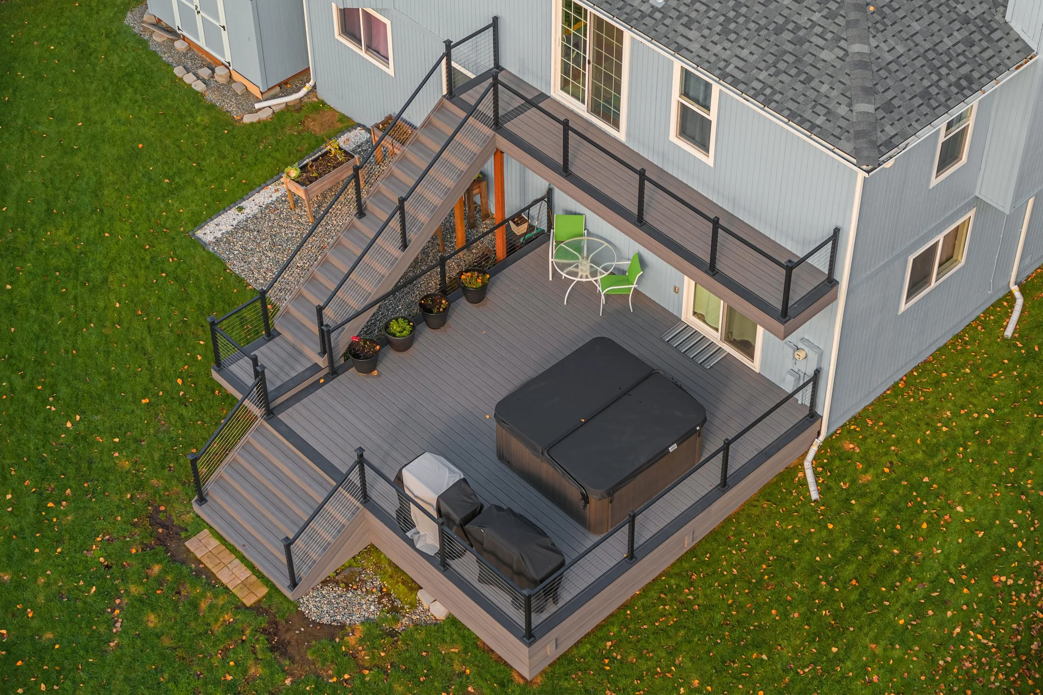 Aerial view of a backyard deck attached to a light gray house with a dark gray roof. The deck has a hot tub, a small round table with two green chairs, and various potted plants along the side. There are stairs leading down from the deck and black sa