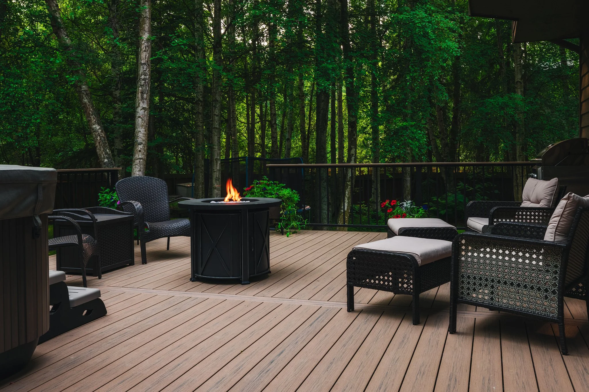 A wcomposite deck outdoor patio with black wicker chairs and a fire pit in the center, surrounded by lush green trees in the background.