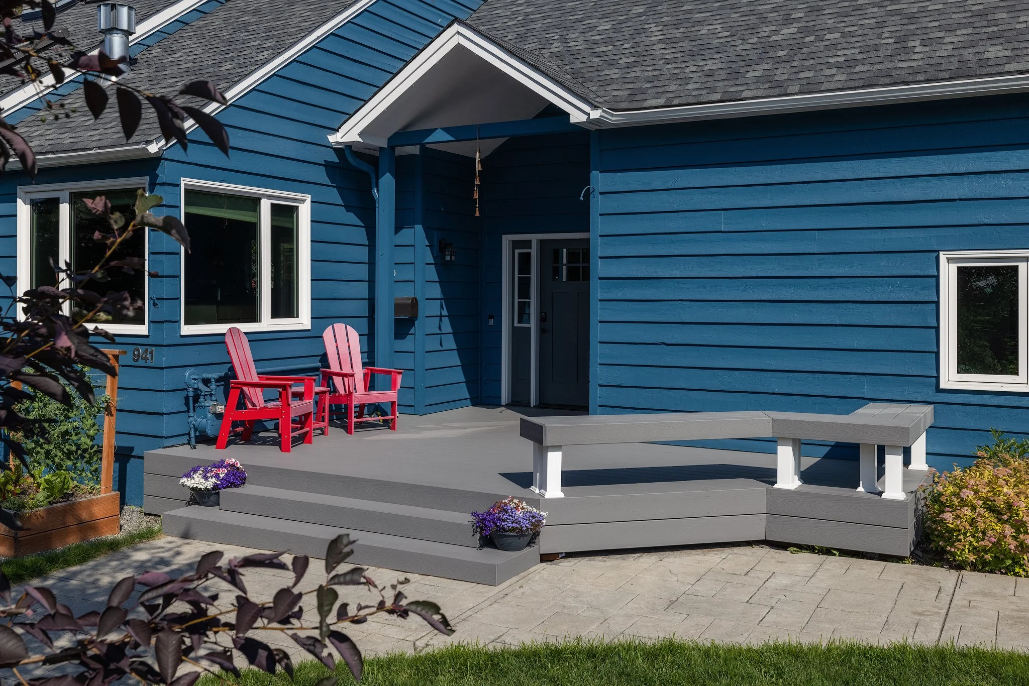 Blue house with a front porch, two red chairs, gray steps, potted flowers, and windows.