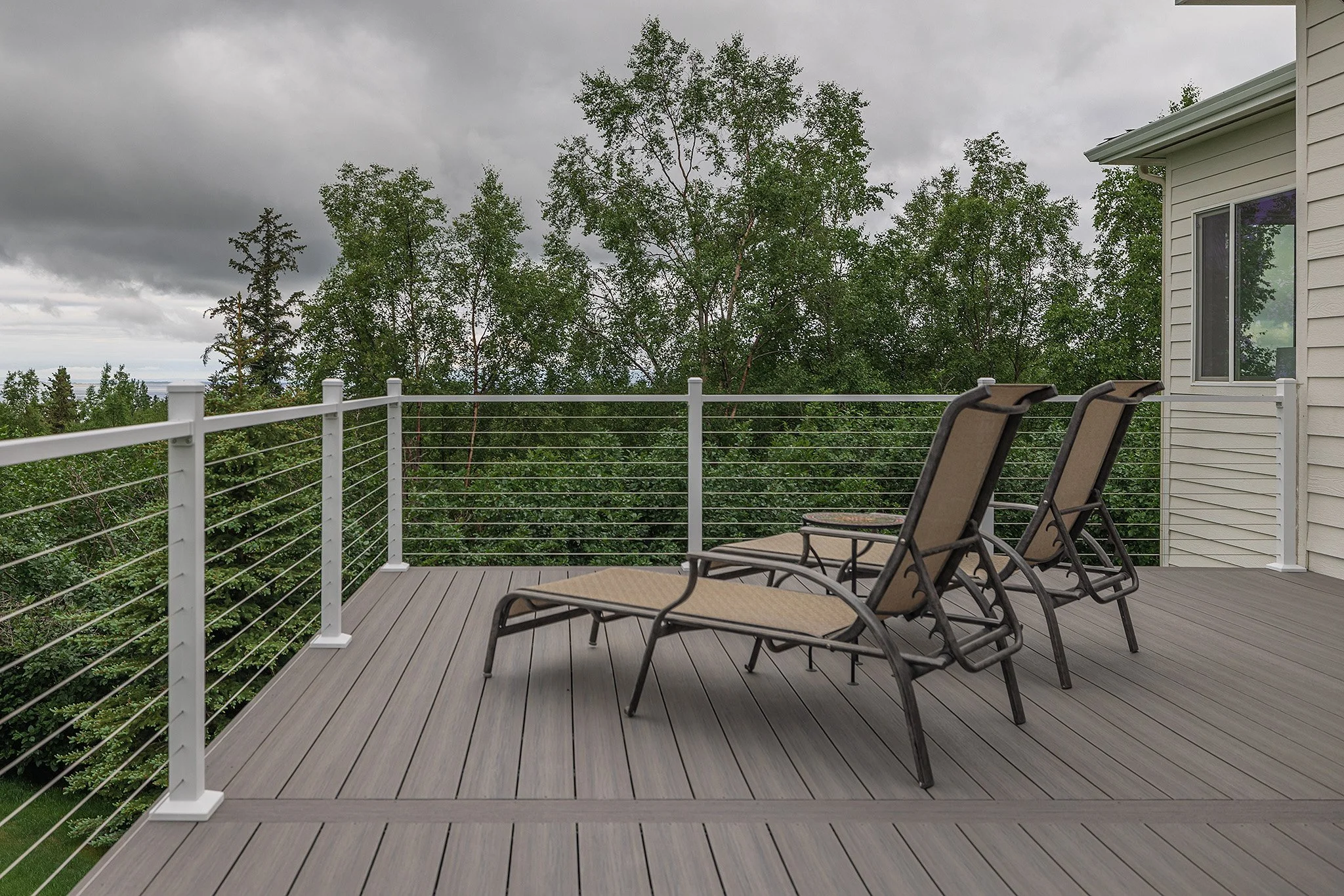 A spacious outdoor composite deck with two lounge chairs facing a green treetop view under a cloudy sky.