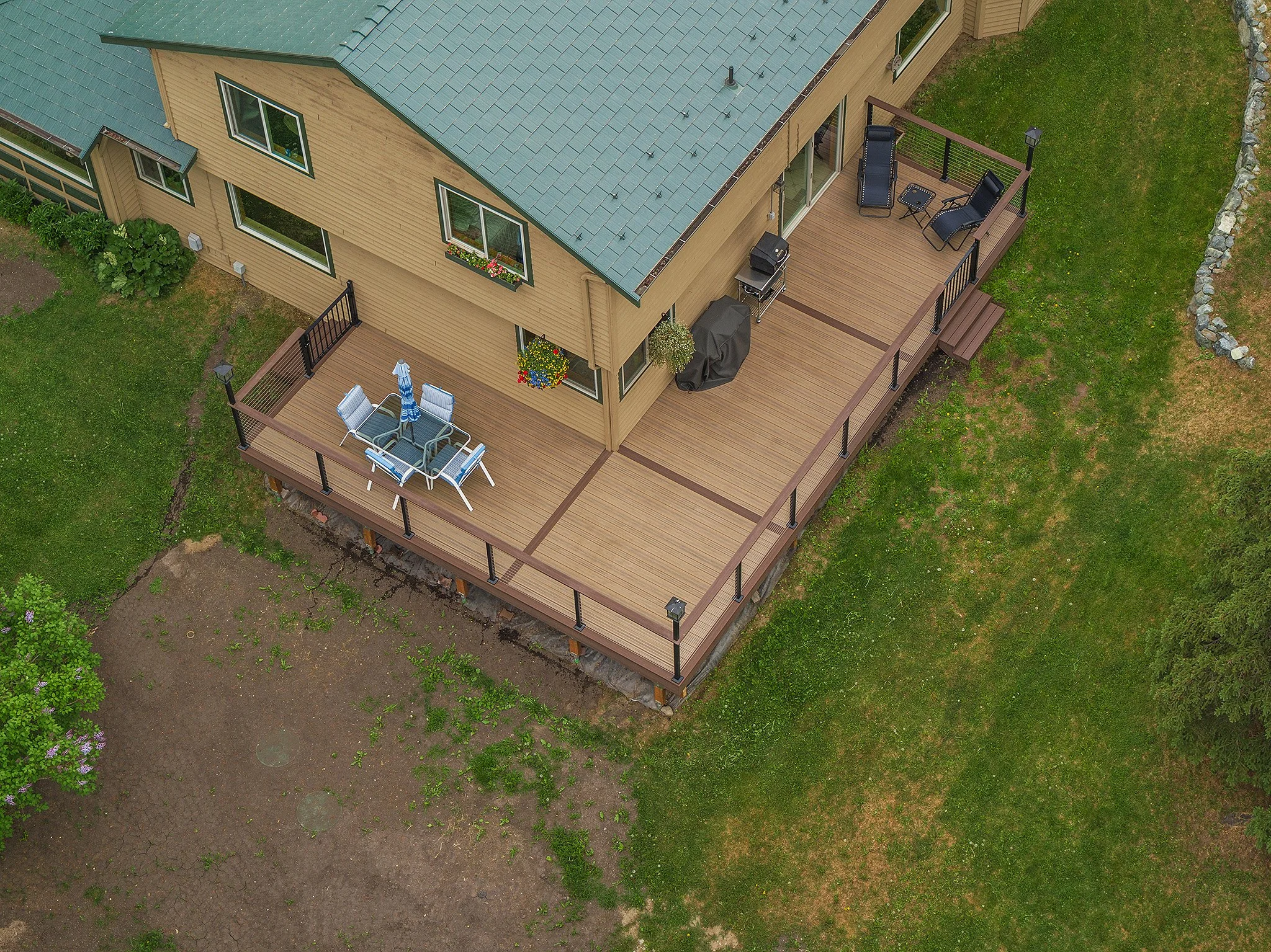 A large composite deck attached to a two-story house with beige siding and a blue roof, featuring outdoor furniture, a grill, and decorative plants amid a grassy yard.