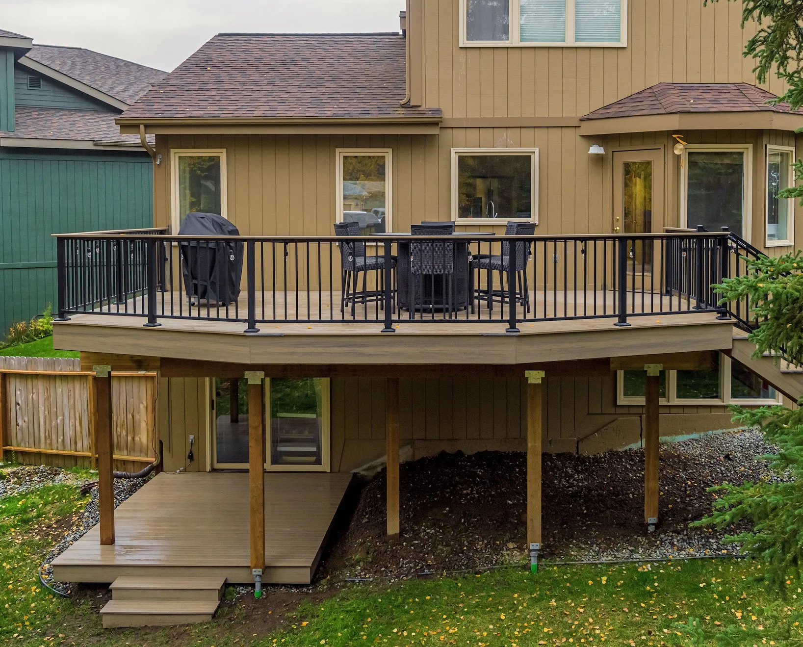 Backyard view of a house with a large composite deck, black railing, outdoor dining table with chairs, and a BBQ grill, with steps leading down to a grassy area and trees.