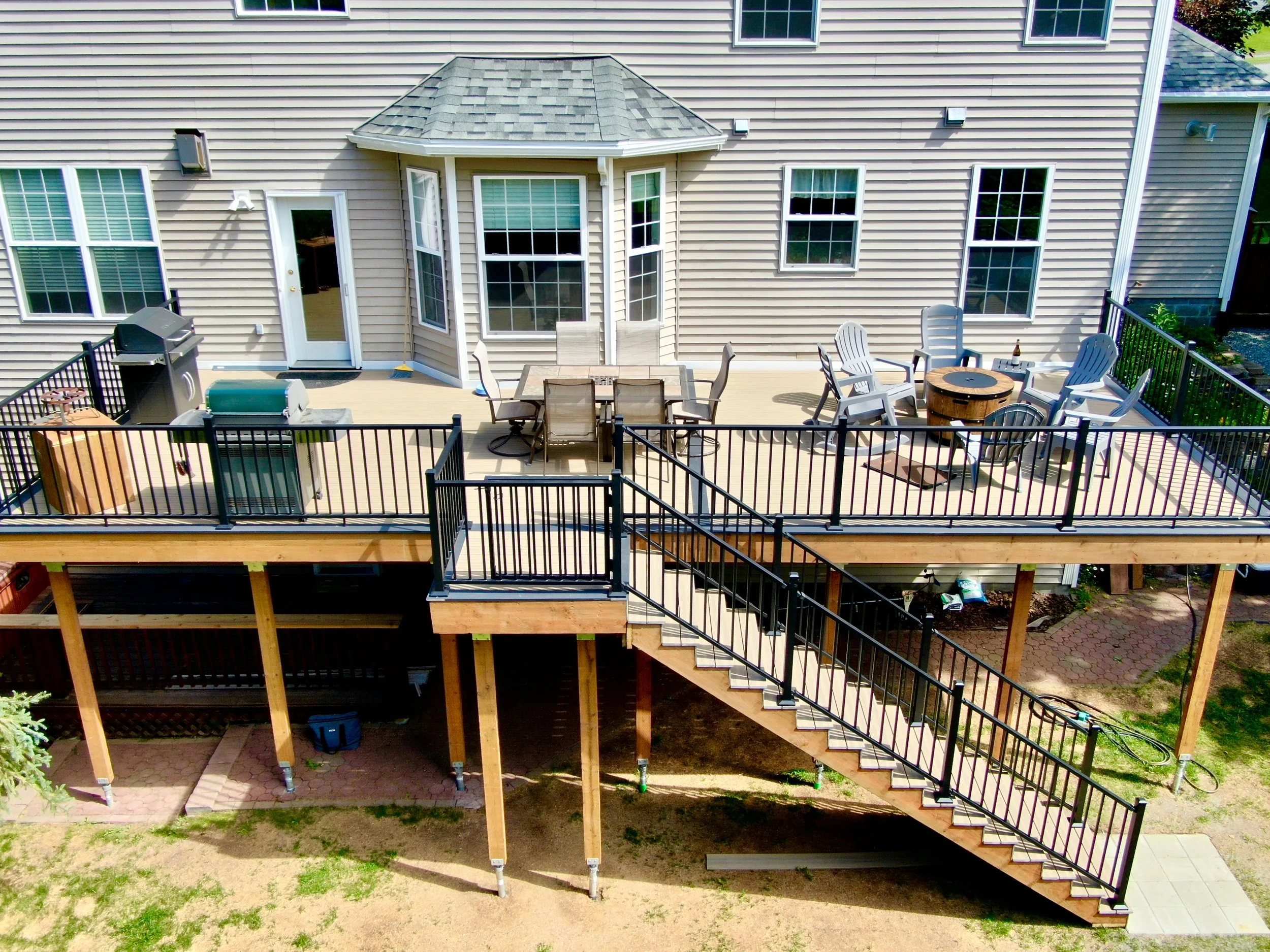 A large elevated backyard deck attached to a house, featuring outdoor furniture, a grill, and a fire pit, with stairs leading down to the yard.