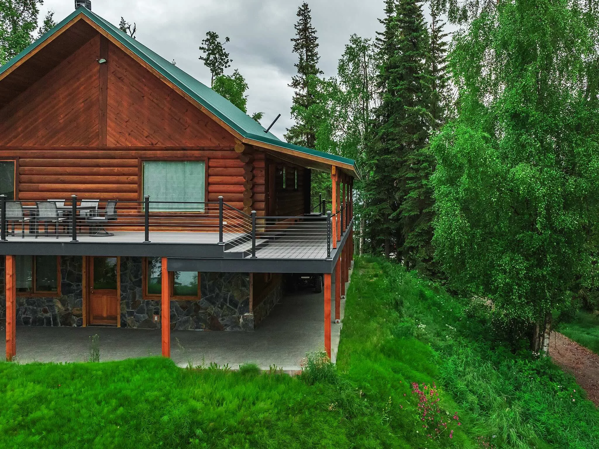 A wooden cabin with a green metal roof and a balcony overlooking a grassy yard and tall trees under a cloudy sky.
