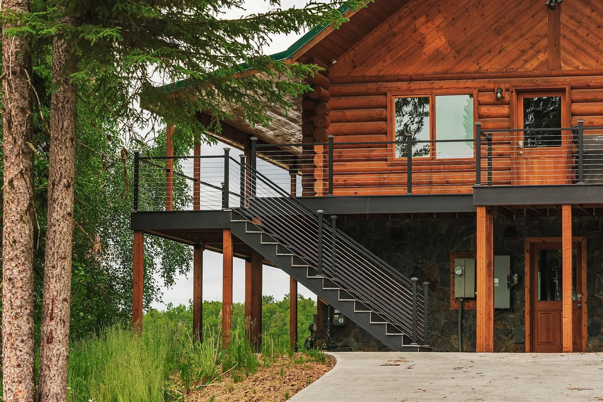 A two-story log cabin with a wooden exterior, metal railing, and a deck surrounded by trees and greenery.