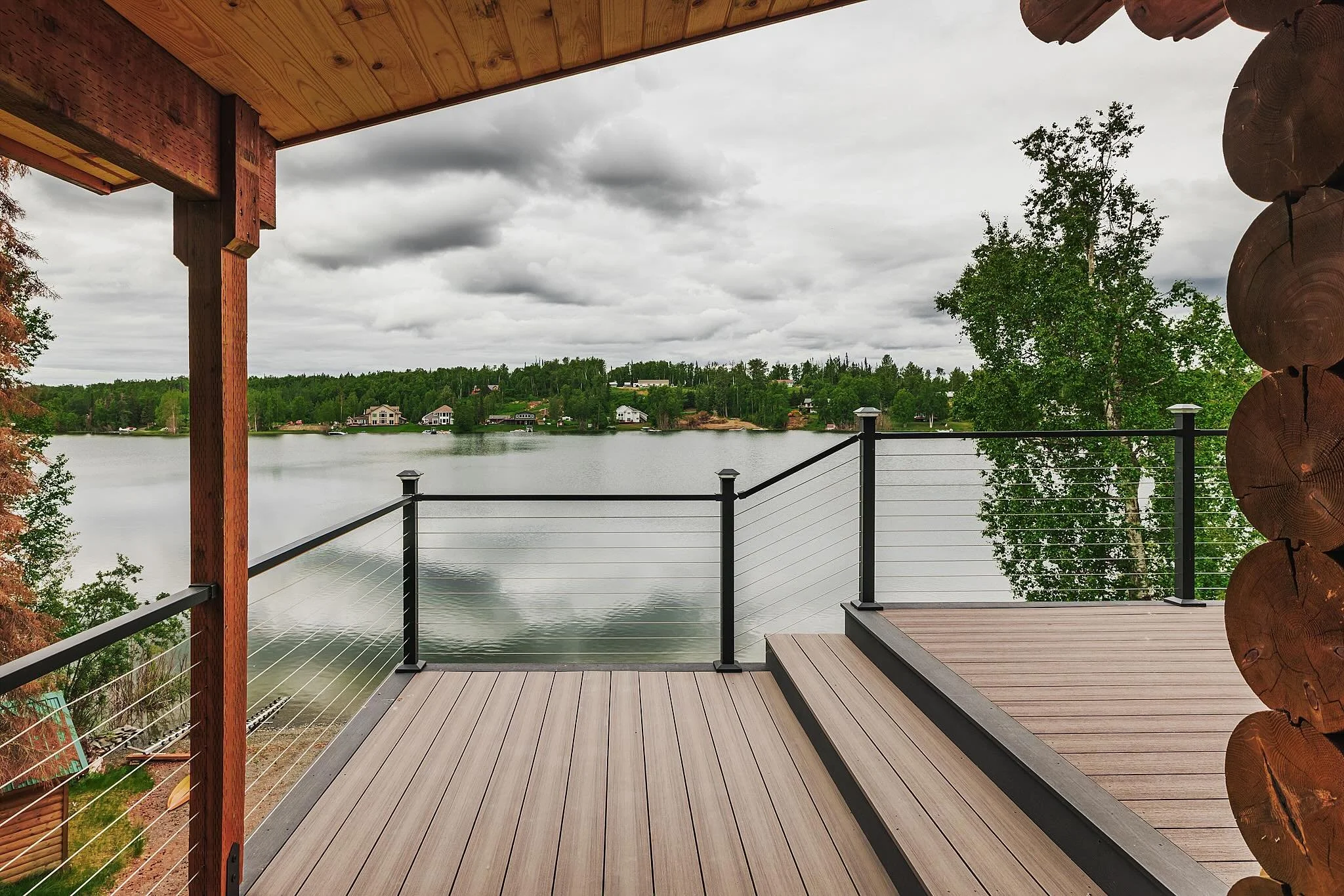 View of a lake with houses in the distance, seen from a composite deck with a black metal railing, under a cloudy sky.