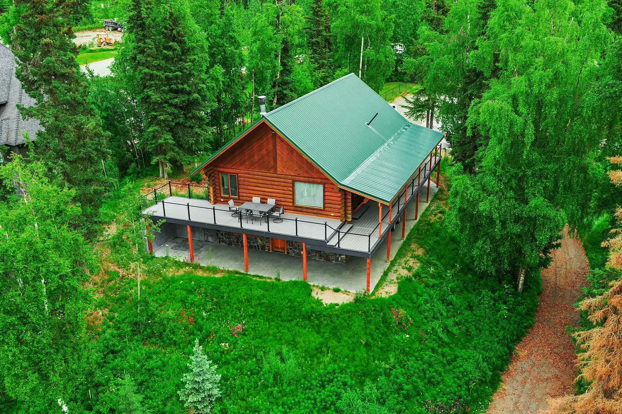 Aerial view of a log cabin with a green metal roof and a large outdoor deck, surrounded by dense green trees.