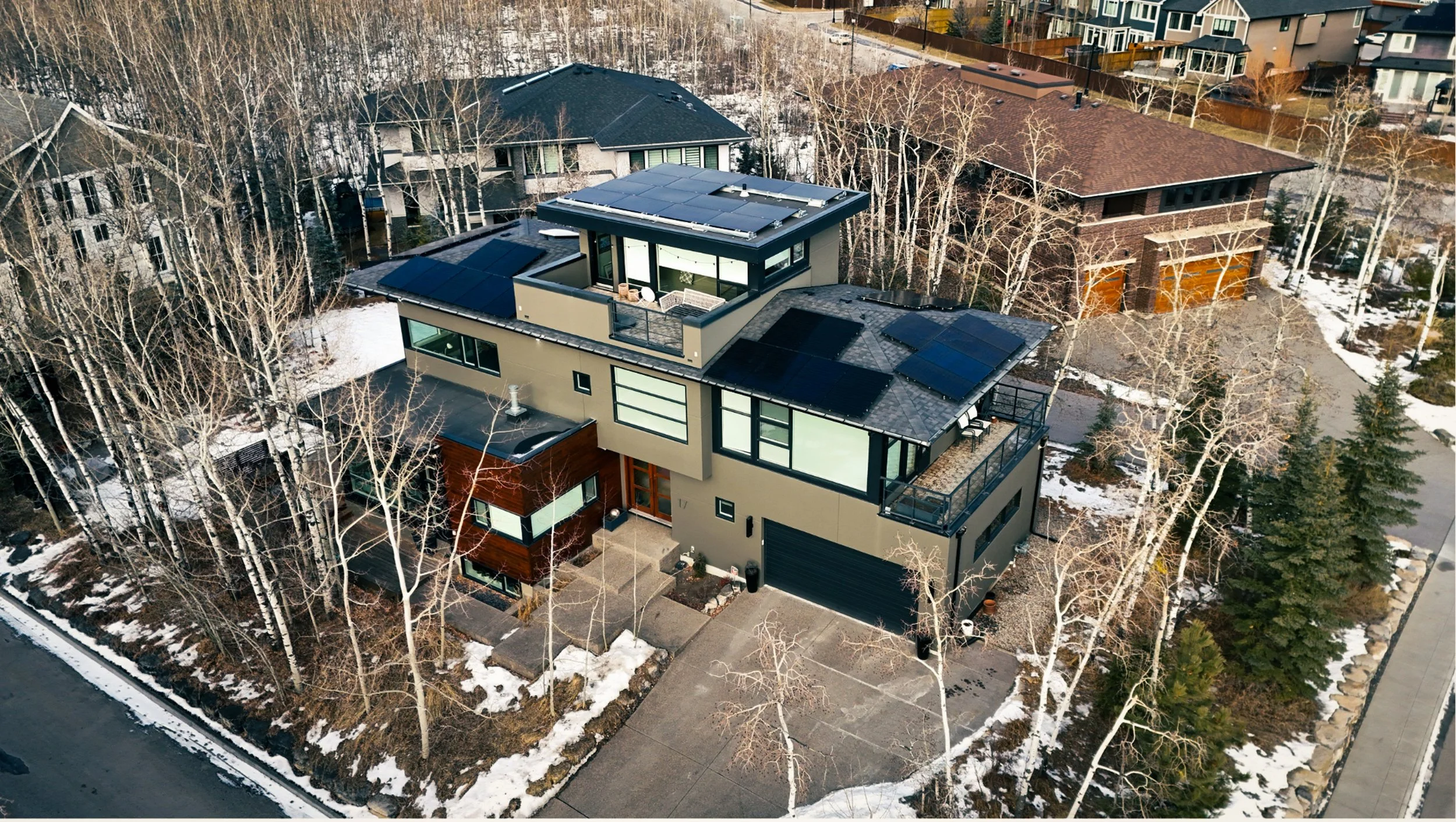 Aerial view of a modern, multi-story house with solar panels on the roof, surrounded by leafless trees and neighboring houses, with patches of snow on the ground.