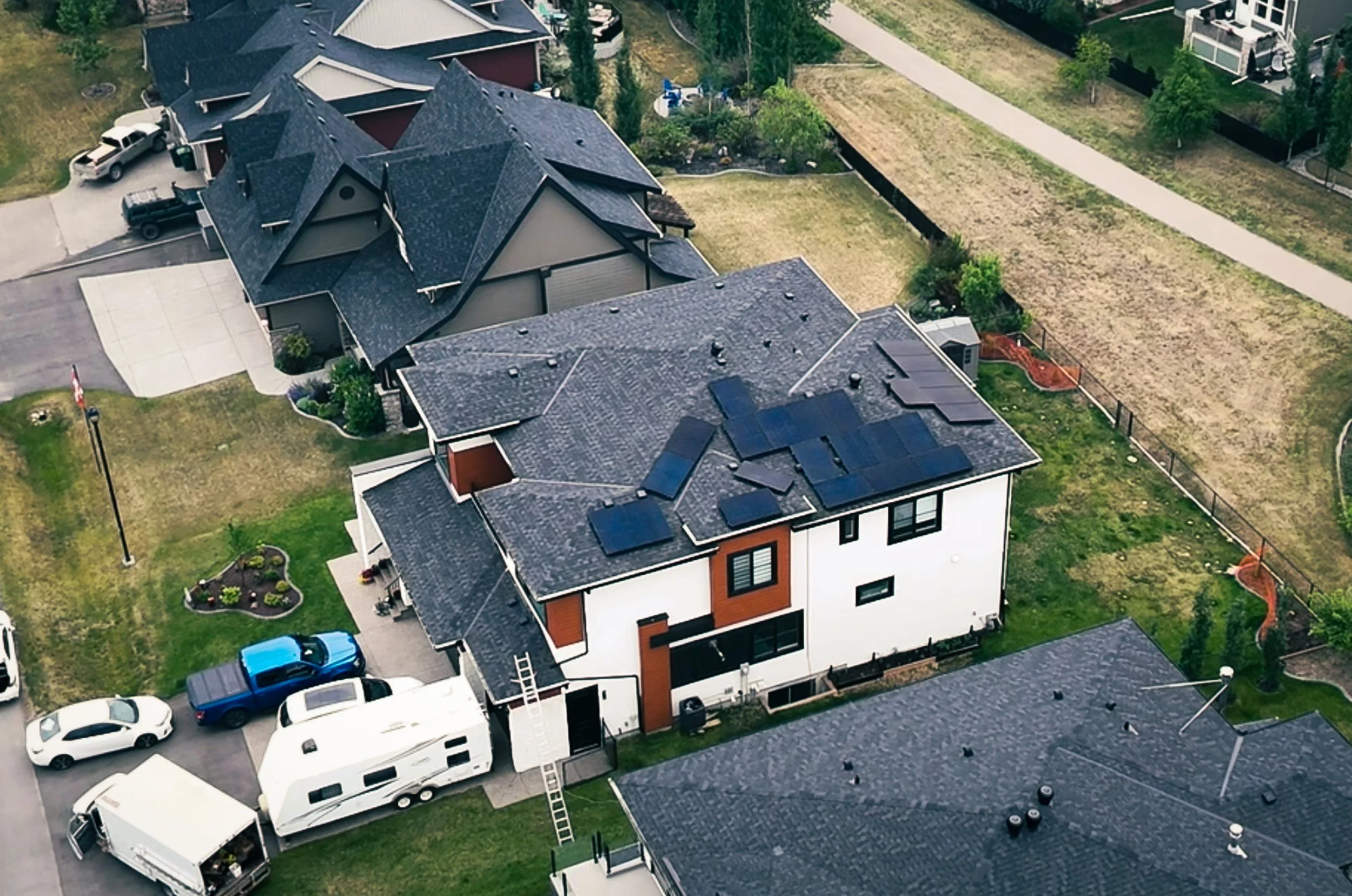 Aerial view of suburban houses with dark roofs, some with solar panels, green lawns, parked cars, and a large vehicle and RV in driveway.