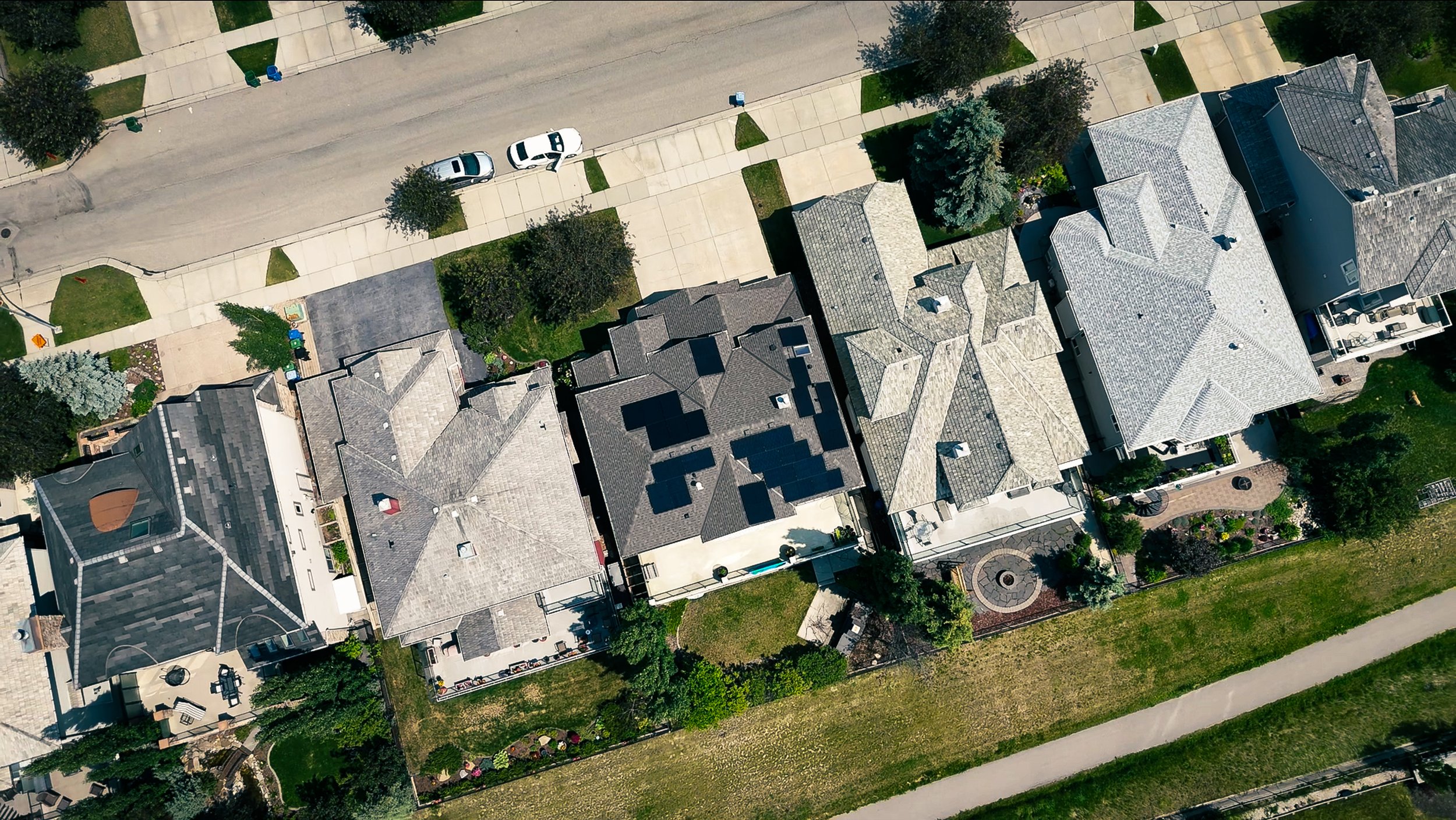 An aerial view of a neighborhood showing houses with various roofing styles, a street with parked cars, sidewalks, and green lawns and trees.