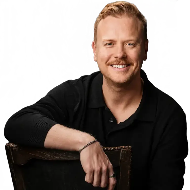 A man with blond hair and a beard, smiling, wearing a black shirt, sitting with one arm resting on a wooden chair.